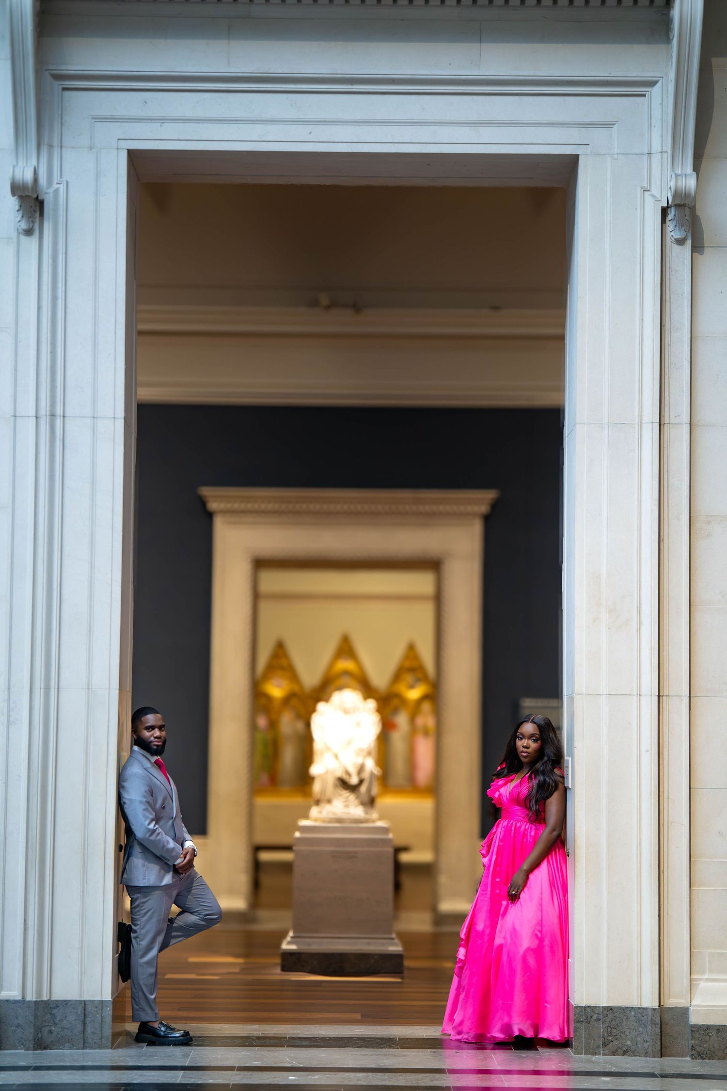 Couple posing in museum doorway, man in gray suit and woman in pink gown, with golden sculpture behind them.