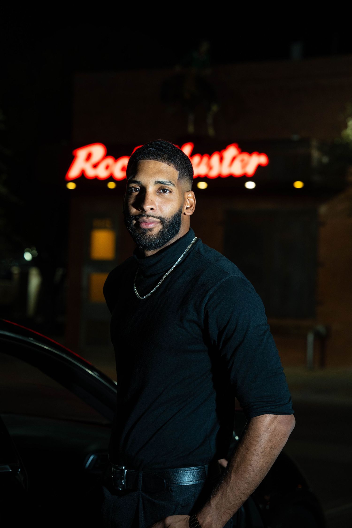A person wearing a black shirt stands in a dimly lit parking lot at night with red neon signs visible in the background.