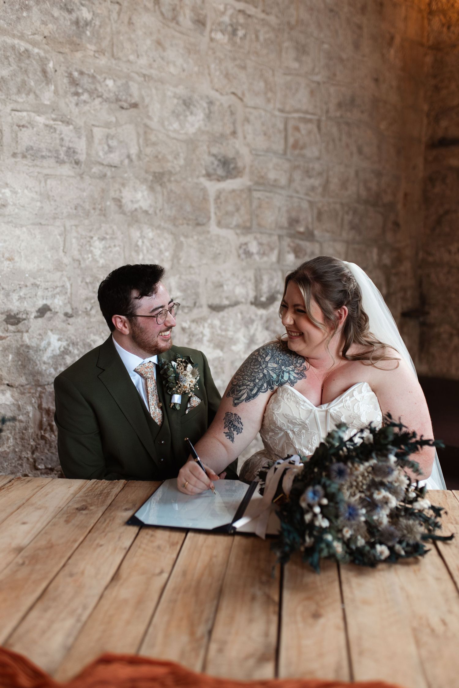 A romantic wedding couple shares a tender moment while sitting at a rustic wooden table against a stone wall.