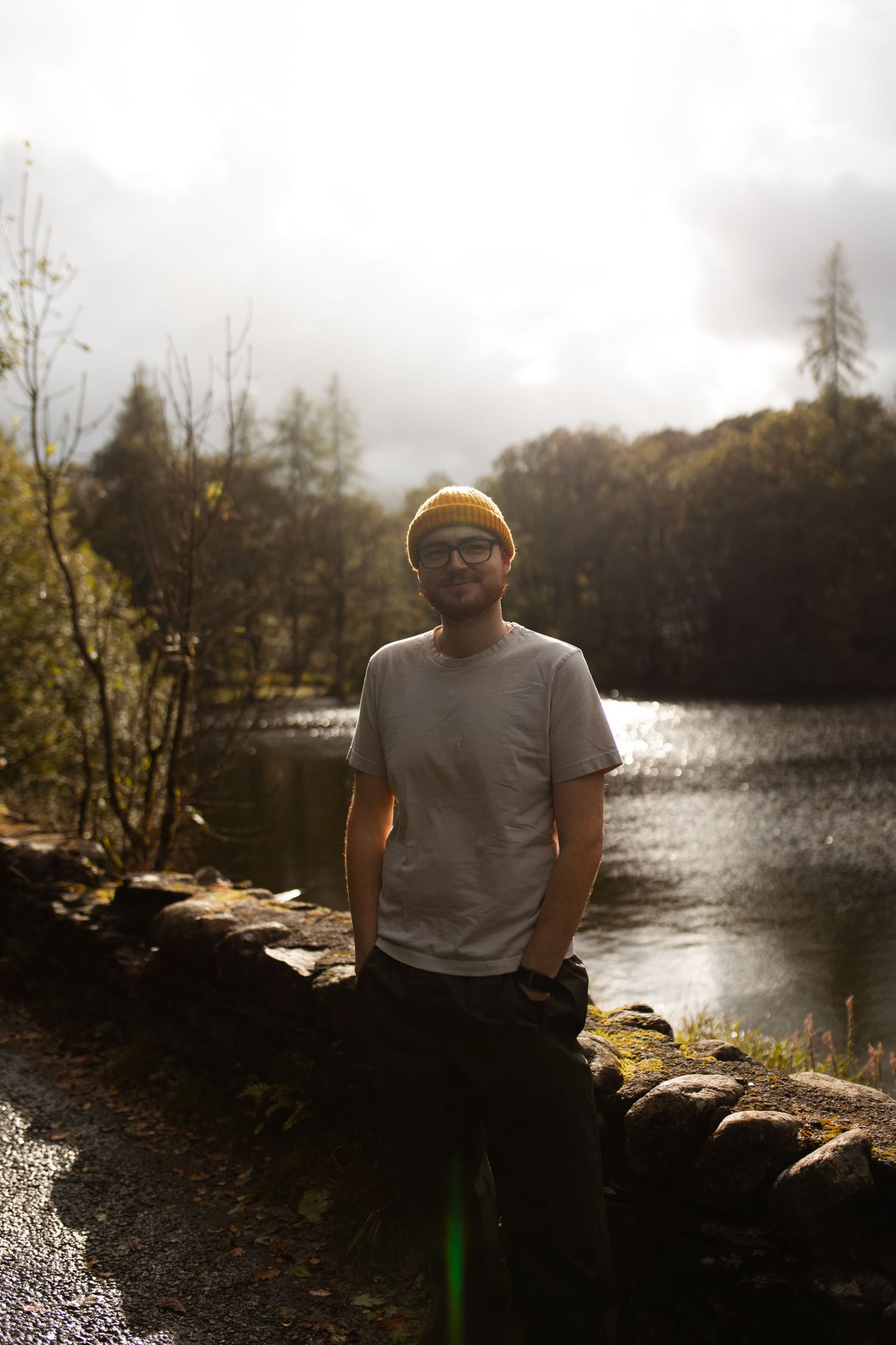 A person sits on a stone wall beside a tranquil lake during autumn with trees and mist in the background.