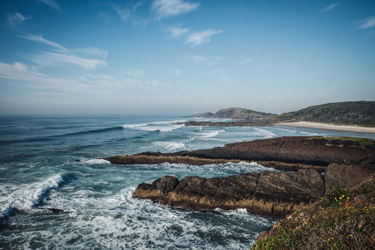 Dramatic coastal landscape with rocky cliffs and crashing waves under a blue sky.