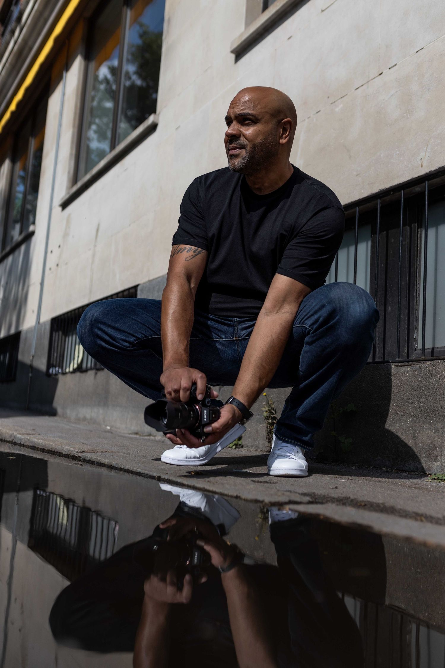 Person in black t-shirt and jeans crouches on sidewalk with reflection in puddle during urban photoshoot.