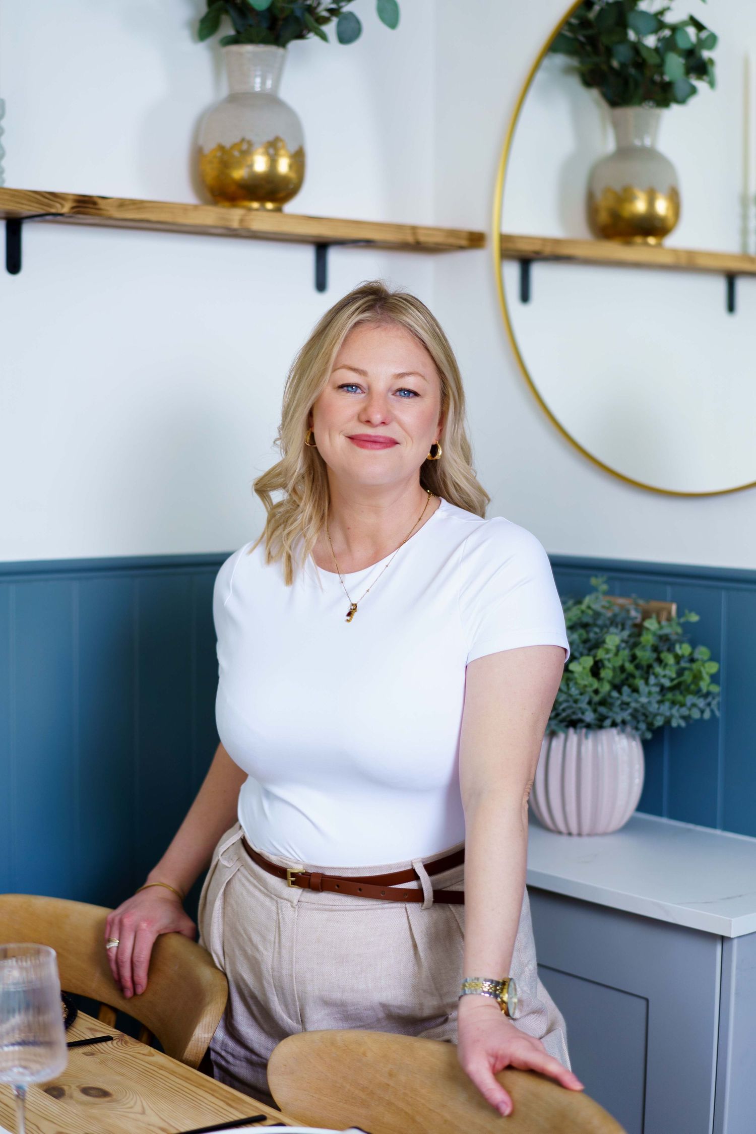 Smiling blonde woman in a white t-shirt and beige trousers stands beside wooden chairs in a bright cafe with blue paneling and gold vases.