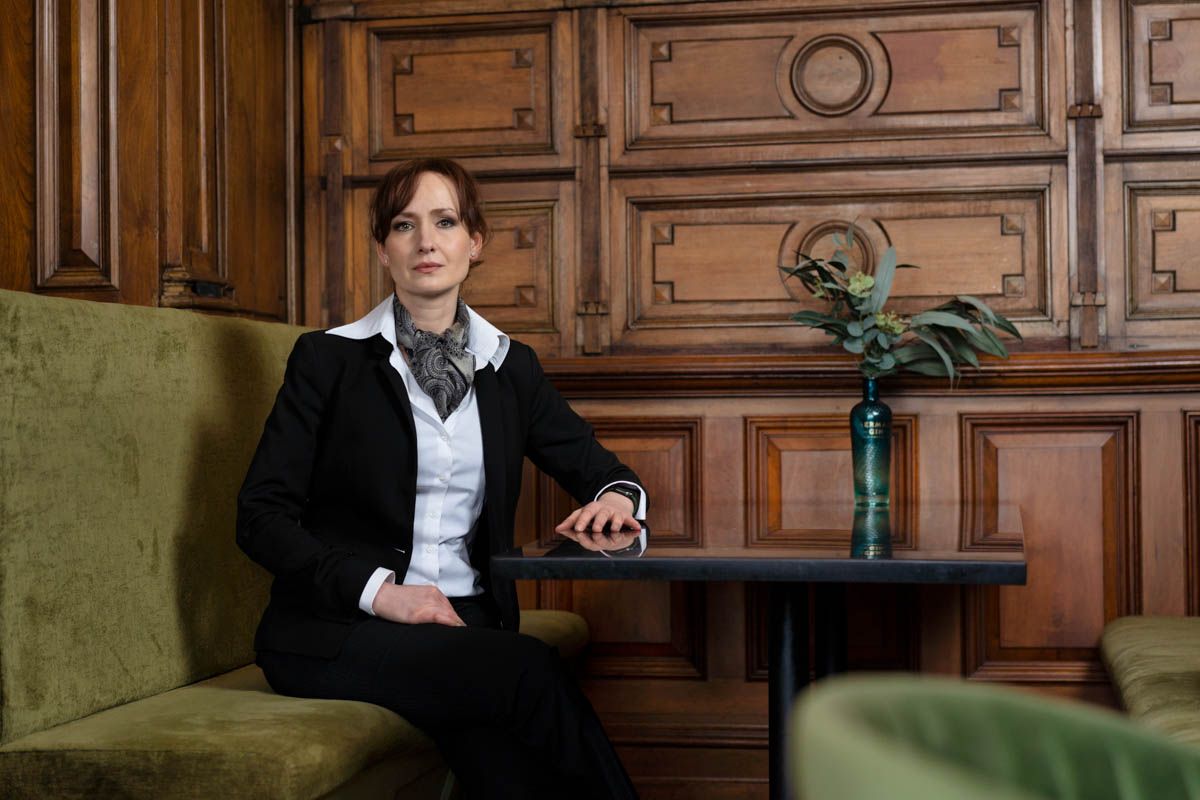 Professional woman in black blazer seated in elegant wood-paneled lounge with green velvet chairs and decorative plant.