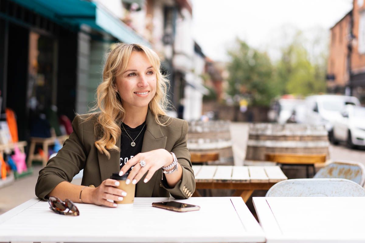Young blonde woman smiling at outdoor cafe table, holding iced coffee with smartphone nearby on sunny day.