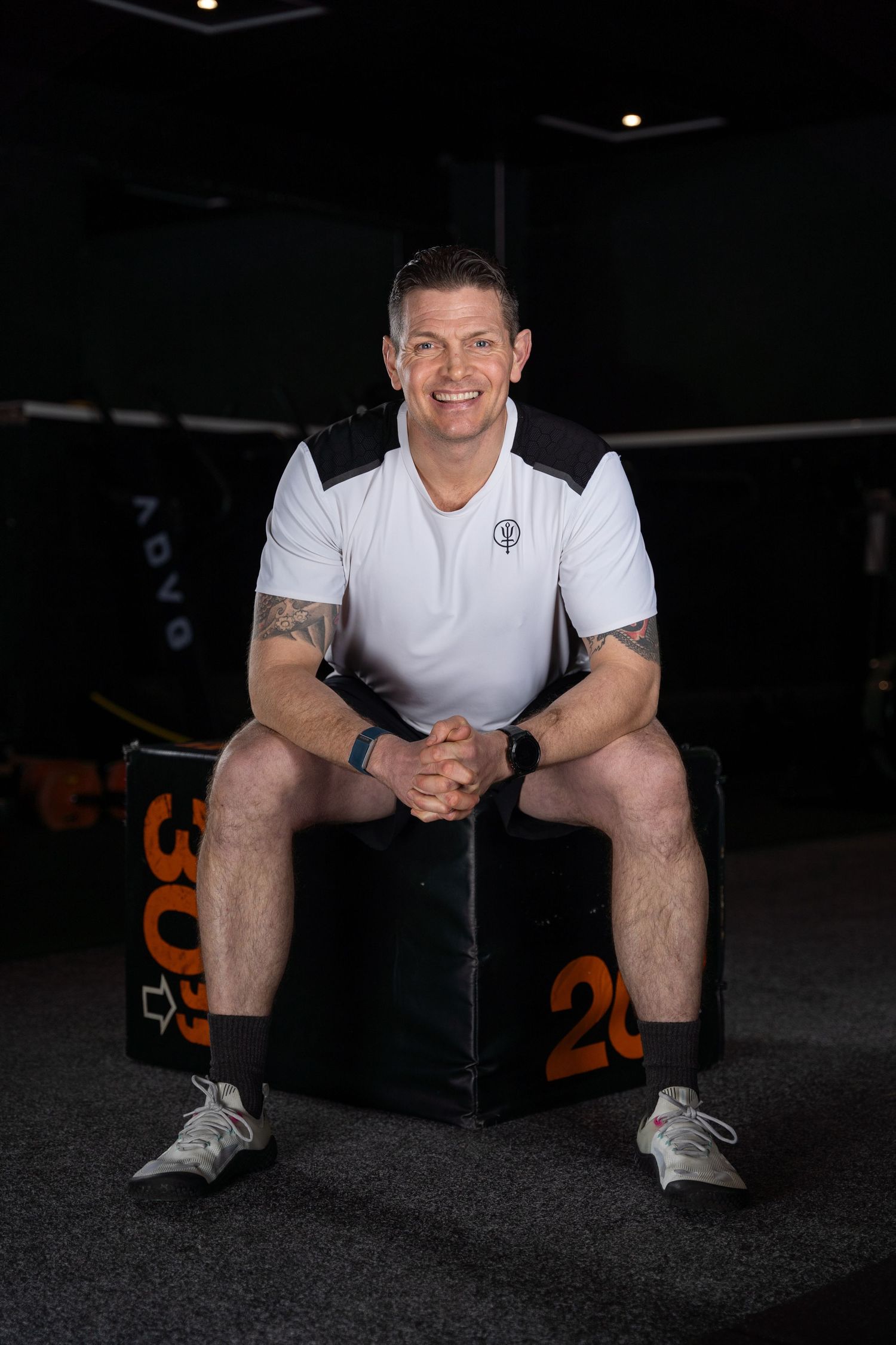 Local fitness coach in white polo shirt sitting on black box in dark gym setting in Fareham during his personal branding photoshoot