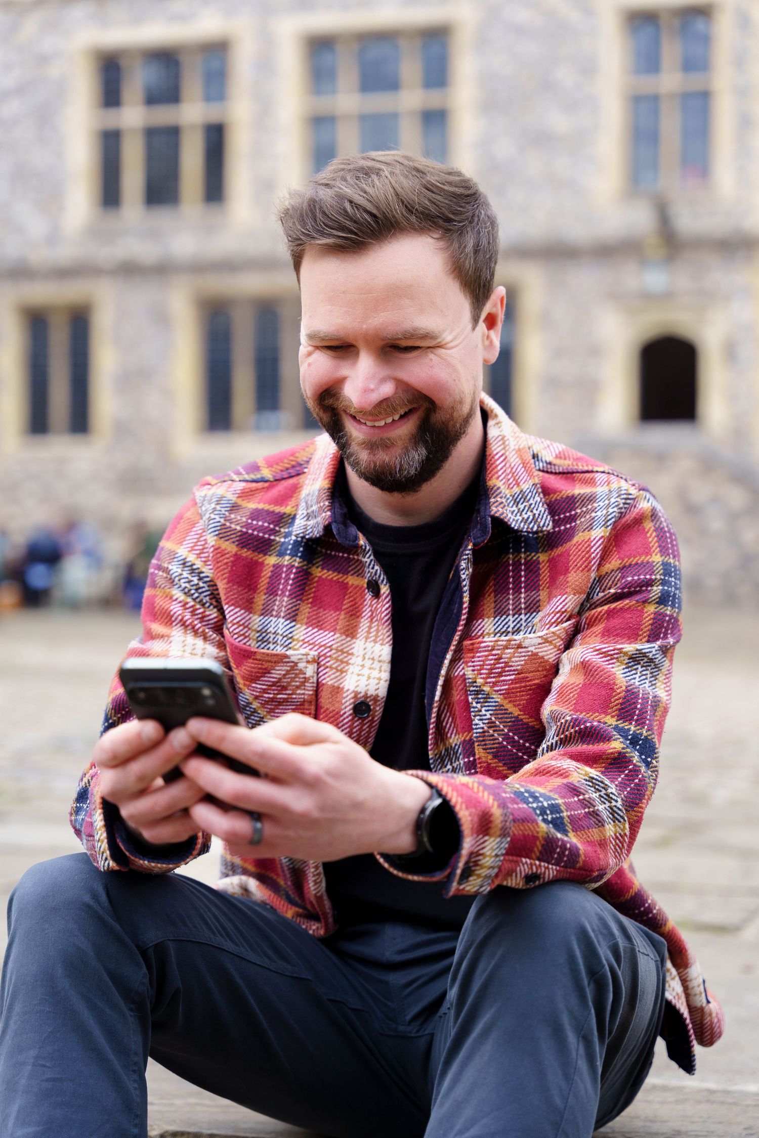 Smiling bearded man in a red plaid flannel shirt sits outdoors on stone steps, looking at his smartphone in front of a historic building.