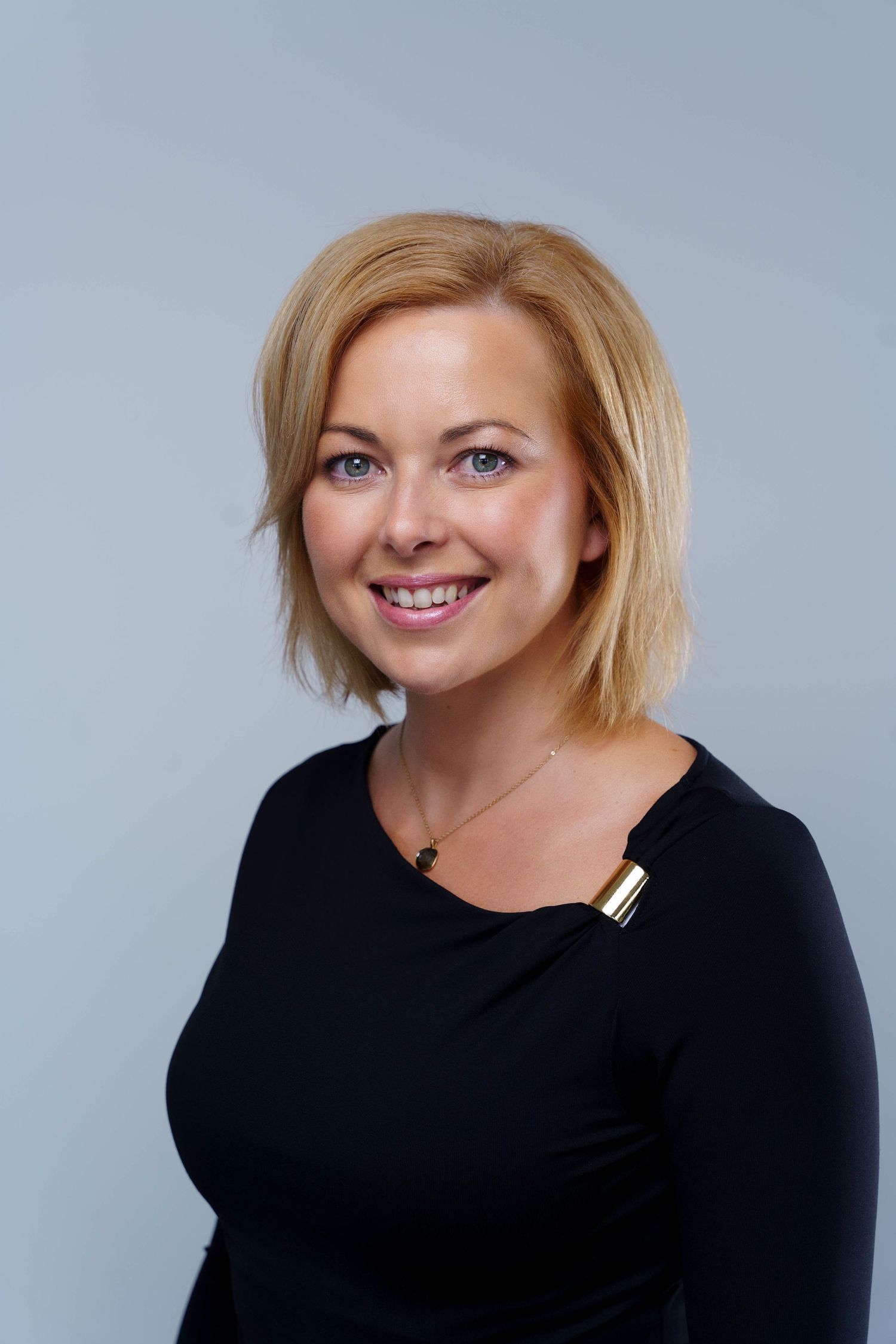 Smiling woman with shoulder-length blonde hair wearing a black top with a gold clasp and pendant necklace in a professional portrait.