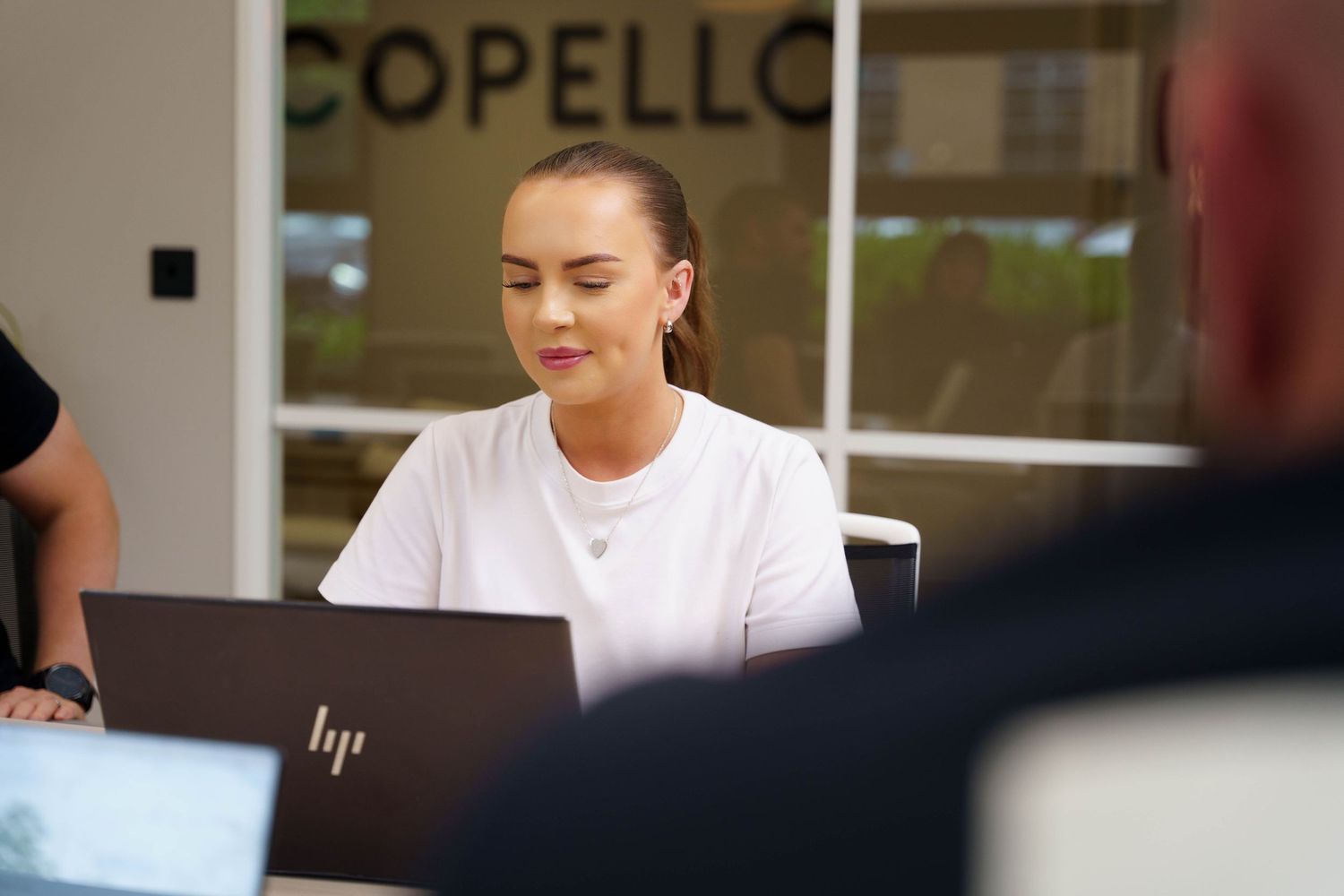 Woman with her hair pulled back works on an HP laptop at a desk in a bright office with a Copello sign in the background.