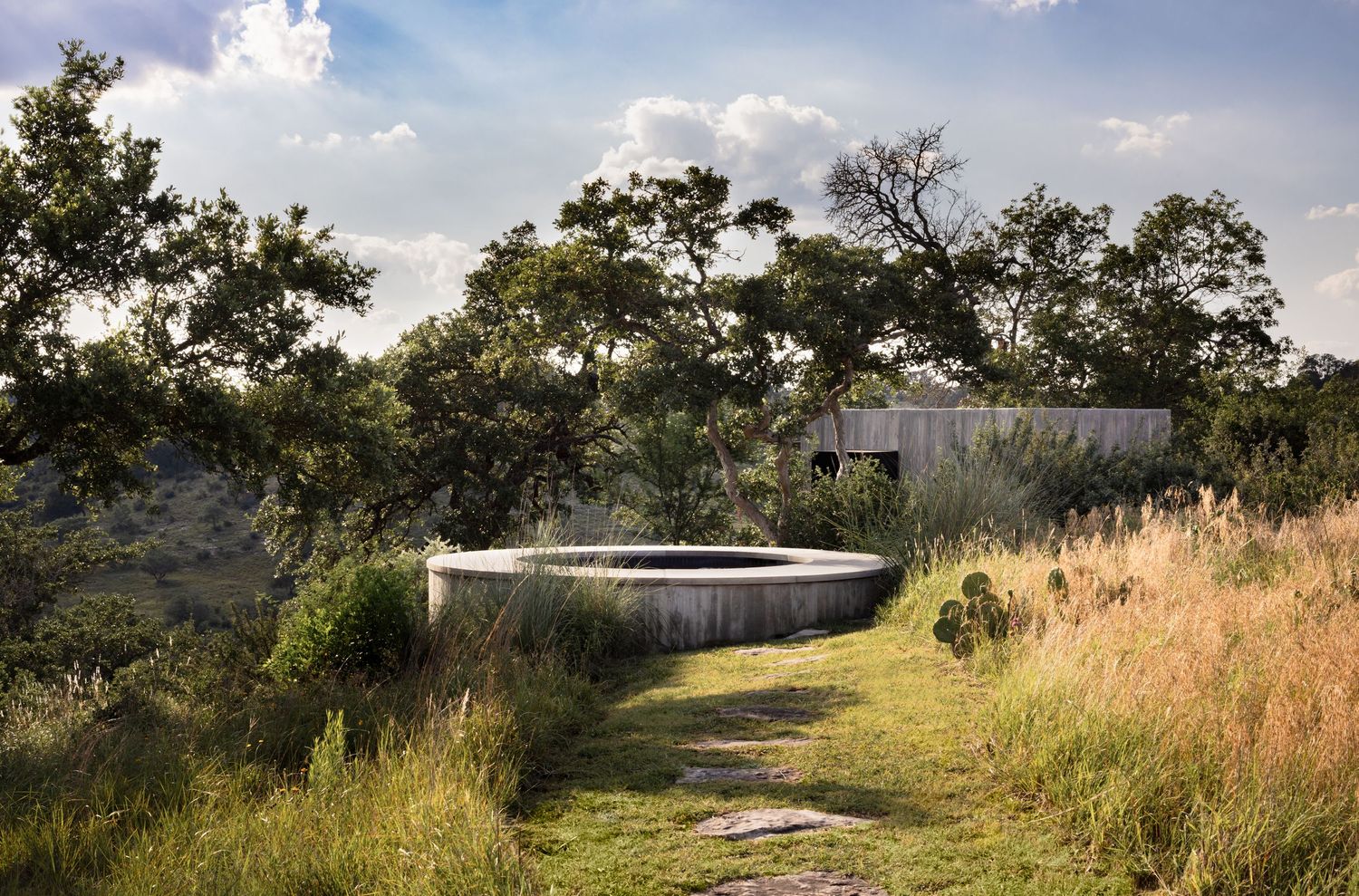 Circular stone water feature surrounded by lush greenery and tall grasses with a rustic building in the background.