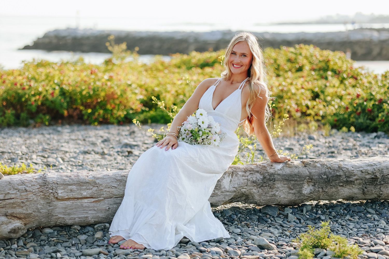 A solo female portrait in a white dress poses gracefully on a rocky beach during golden hour.