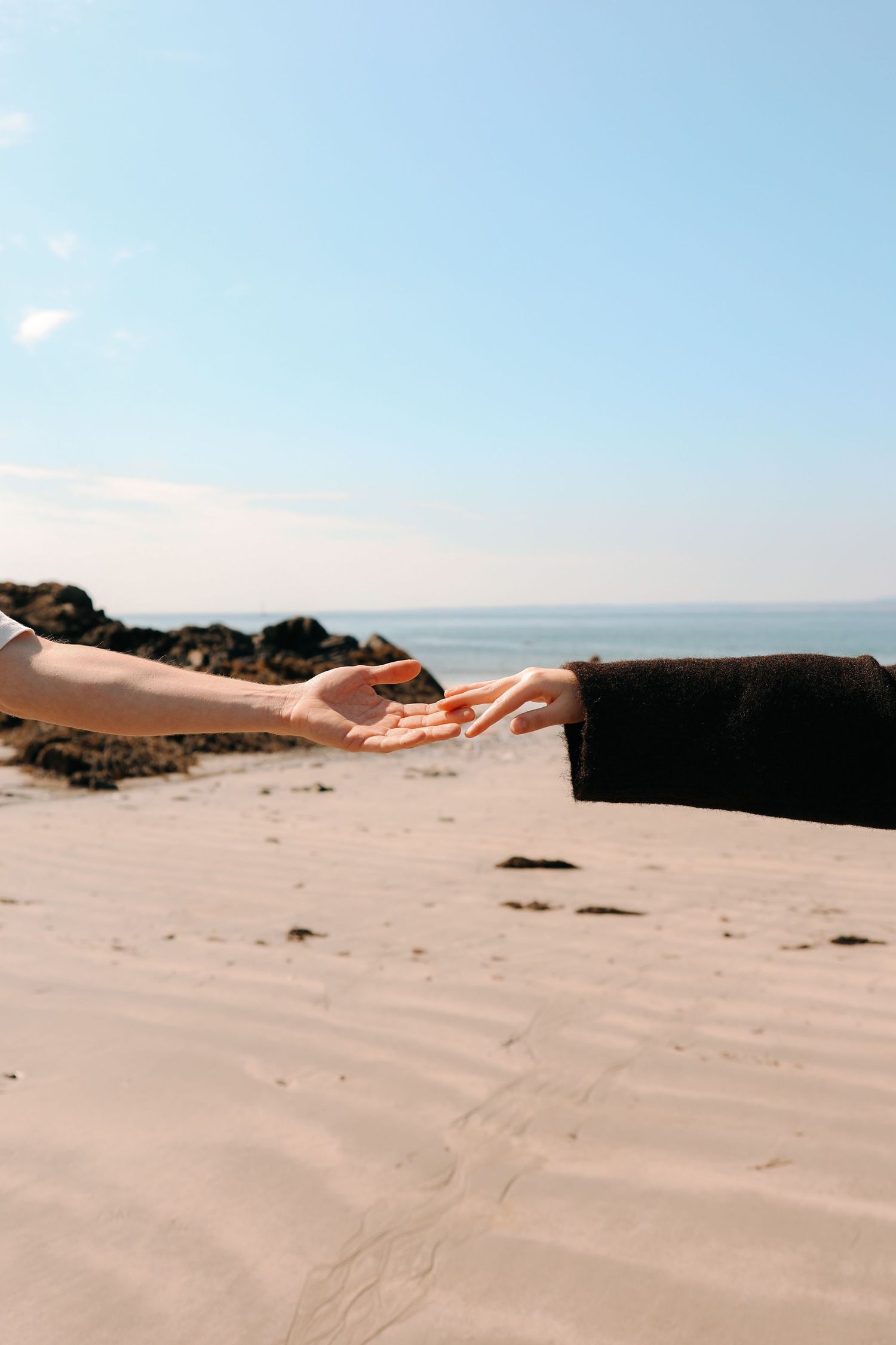 Two hands reaching toward each other on a sandy beach with ocean in background.