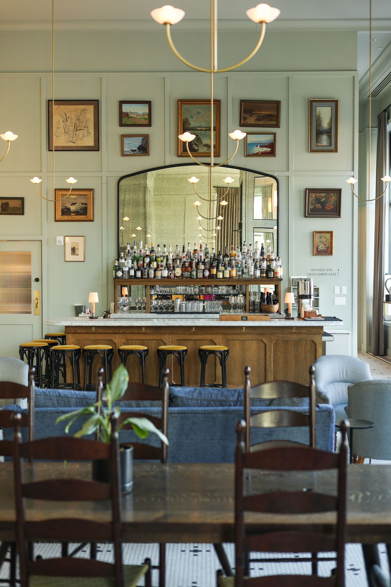Elegant restaurant interior with wooden tables, blue chairs, and a fully stocked bar against white walls with framed artwork.