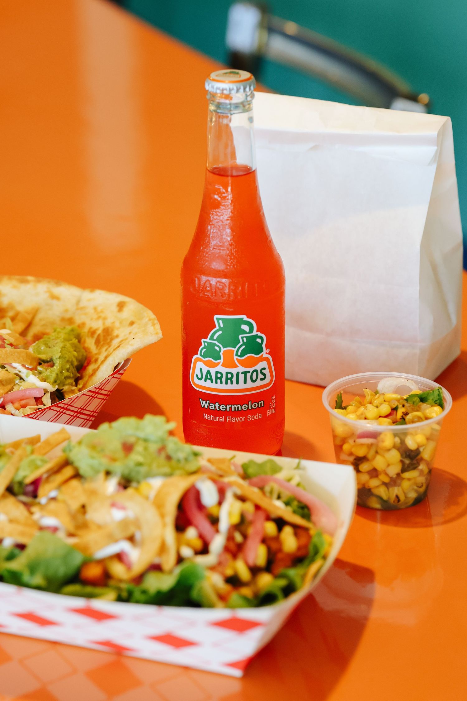 Mexican food plates with tacos and burritos served alongside bright orange Jarritos watermelon soda bottles on orange table.