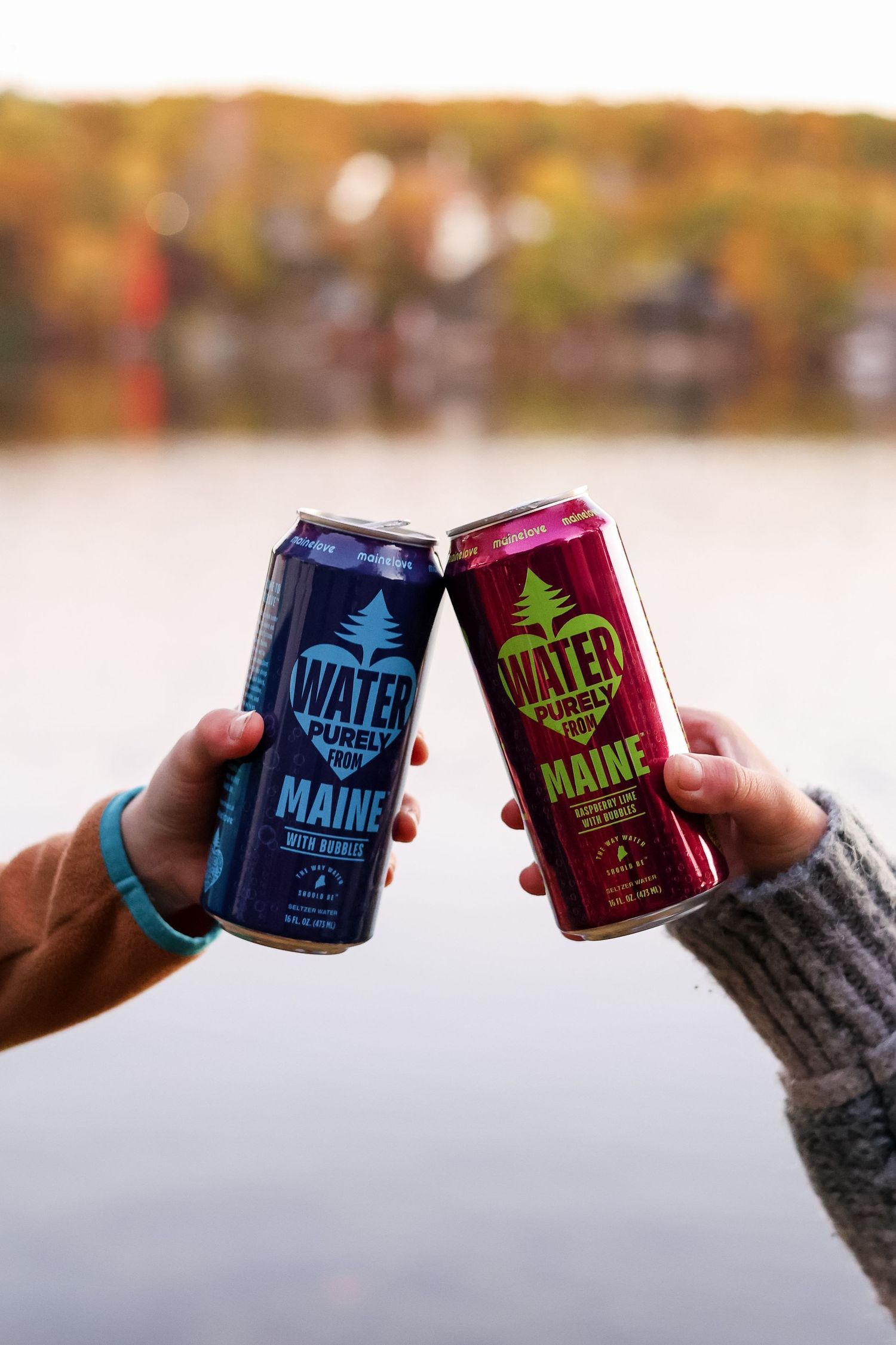 Two hands toast with cans of Water Purely from Maine, one blue and one red, beside a calm lake framed by colorful autumn foliage.