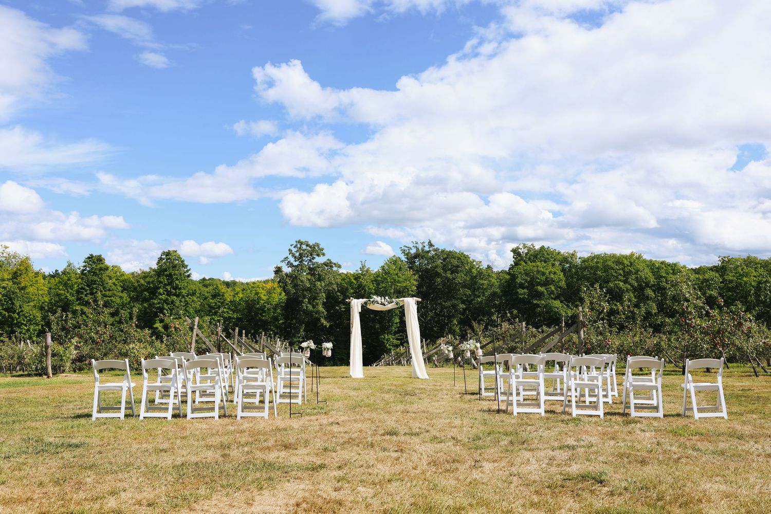 Outdoor wedding ceremony setup with white chairs and arch in a meadow setting.