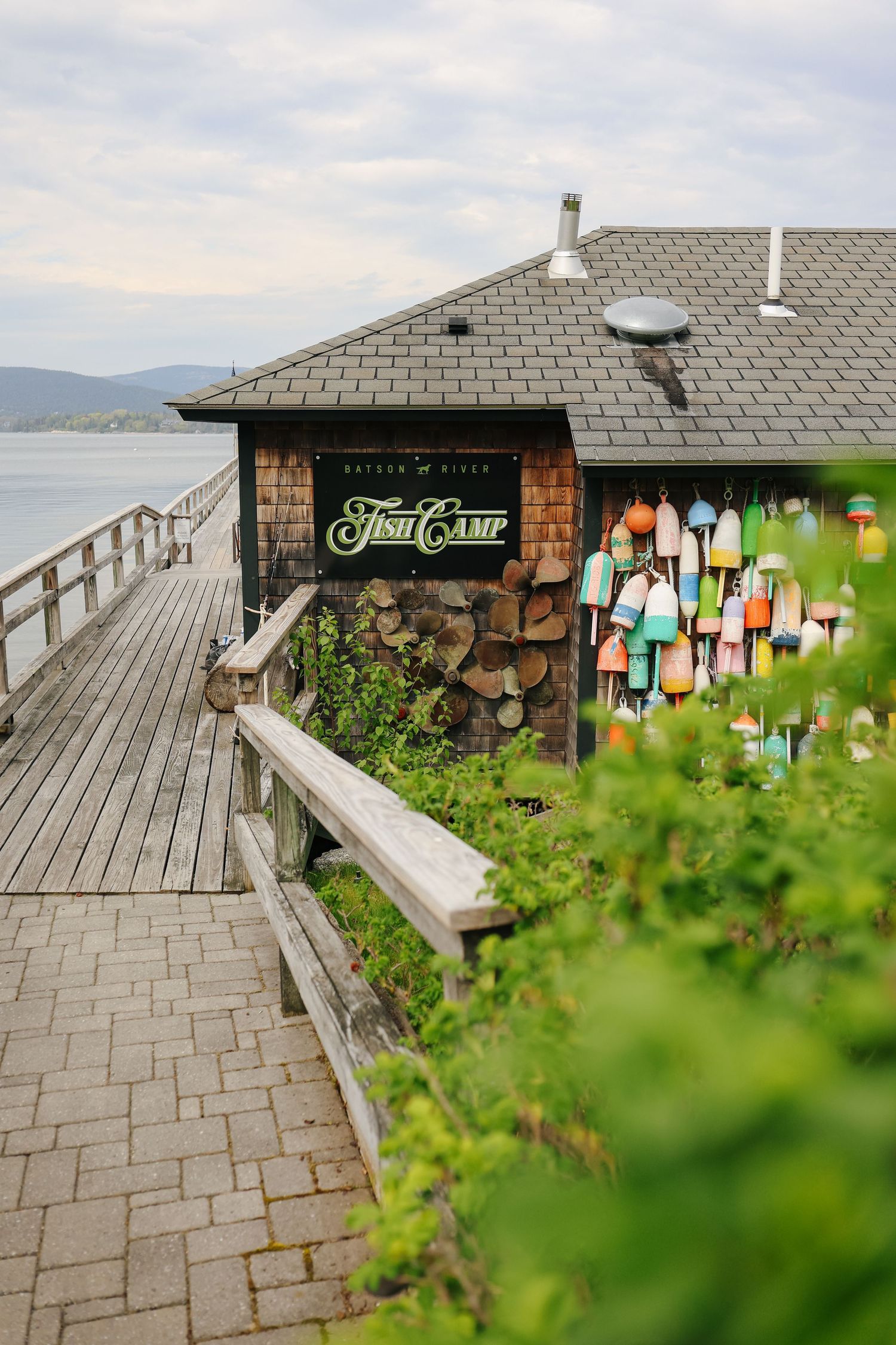 Rustic Fish Camp restaurant on a waterfront boardwalk with colorful buoys and stacked firewood by the sea.