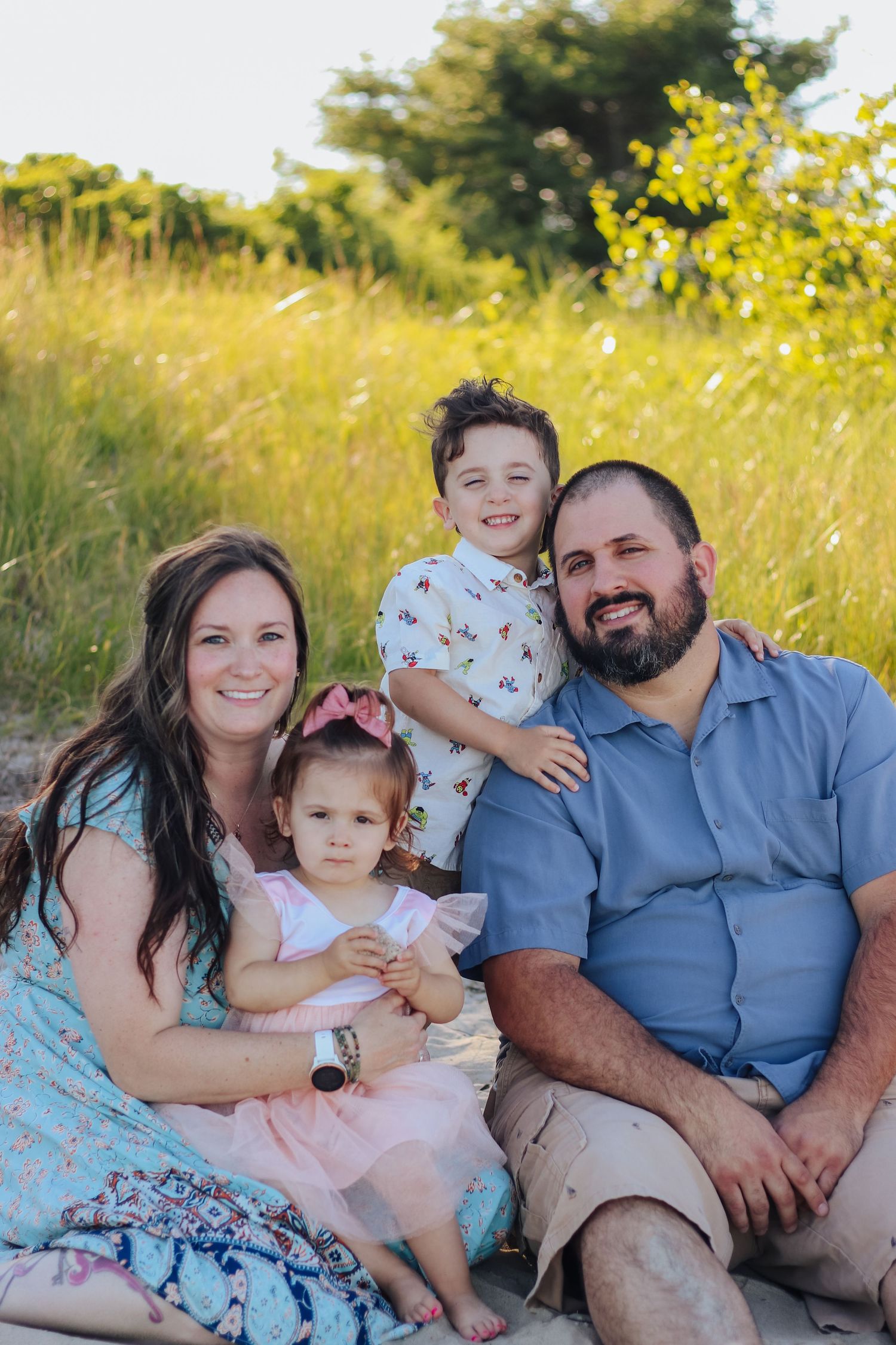Happy family of four posing outdoors in a sunny green field, parents smiling with two young children.