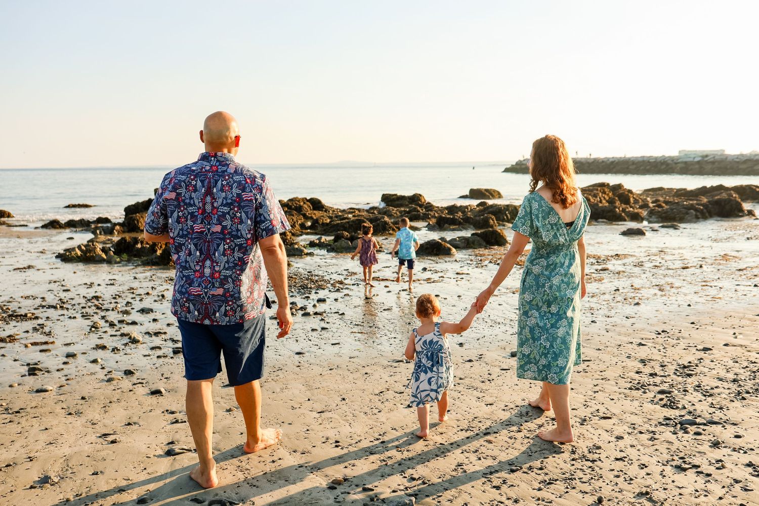 Family walking barefoot on a rocky beach at sunset, parents holding toddler's hands while older child explores the shore.