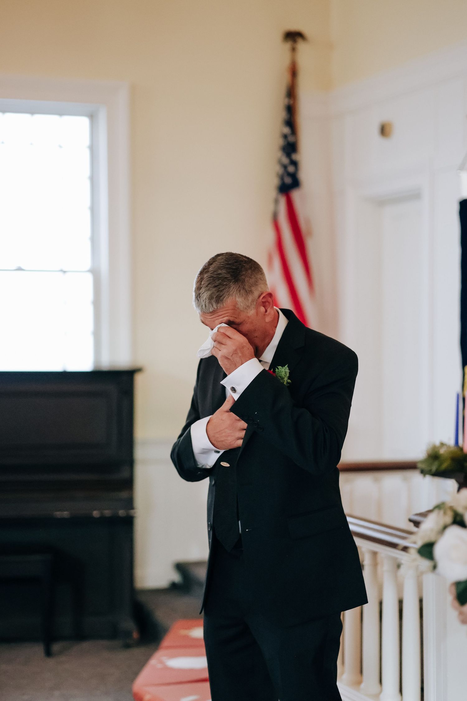 A person in a black suit stands in a white room with an American flag and piano visible in the background.