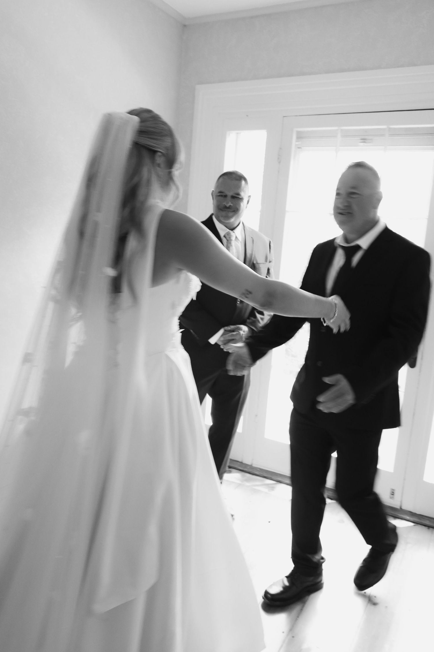Black and white photo of a bride in a white gown and veil greeting two smiling men in suits at a wedding.