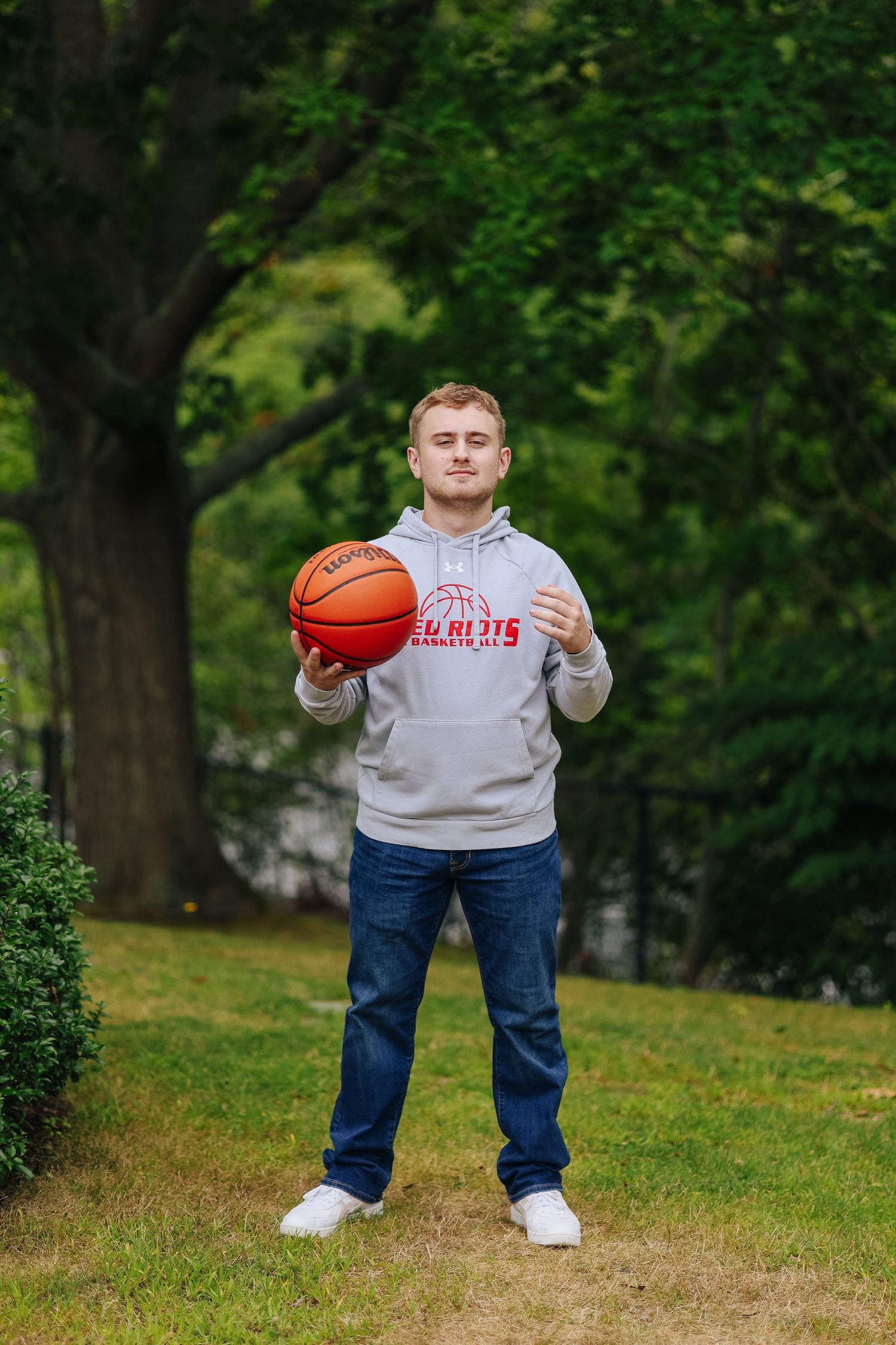 Solo male portrait in grey hoodie and jeans holding basketball outdoors in park setting.