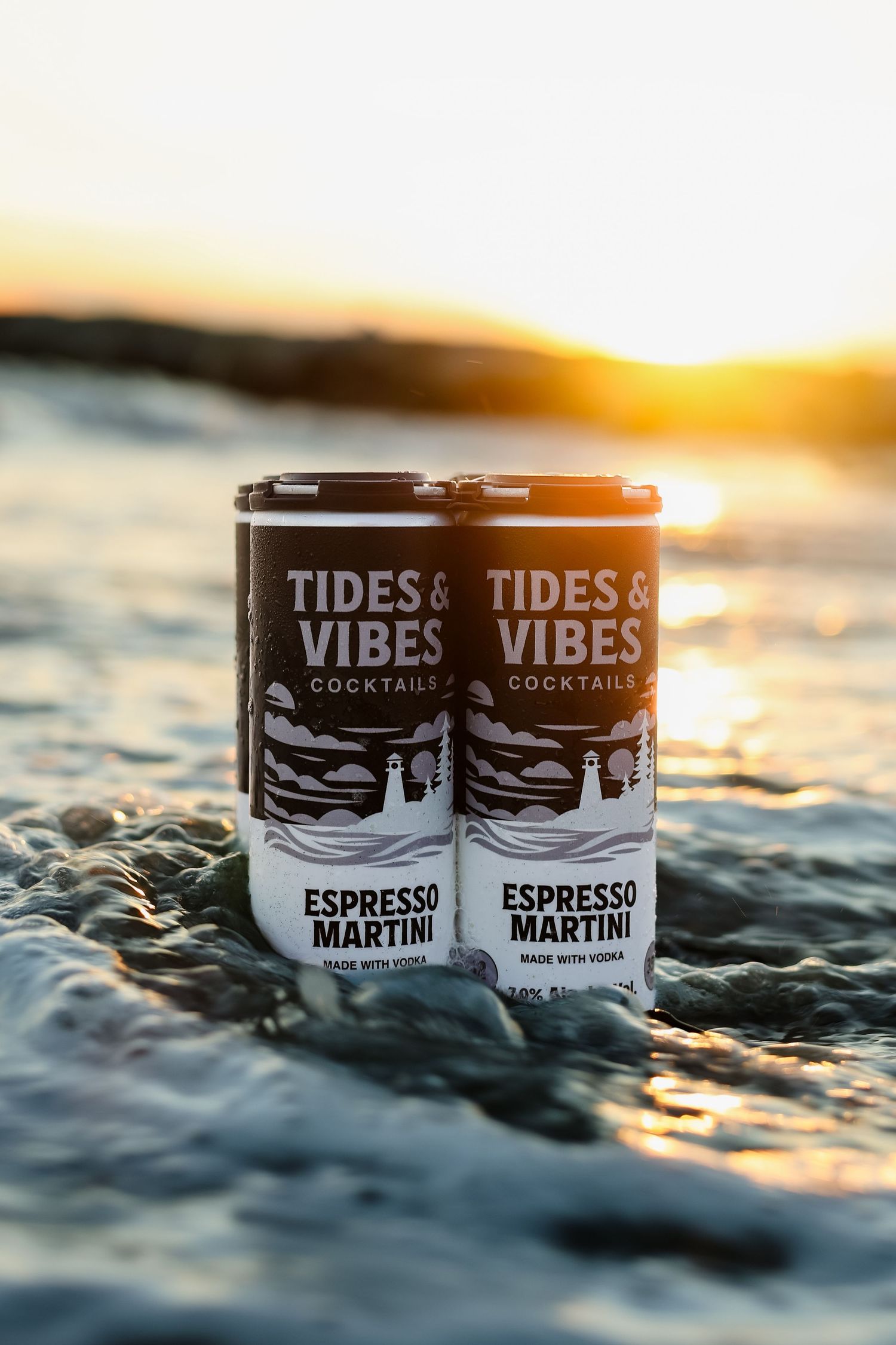 Tides & Vibes Espresso Martini canned cocktails made with vodka sit in shallow ocean water at sunset.