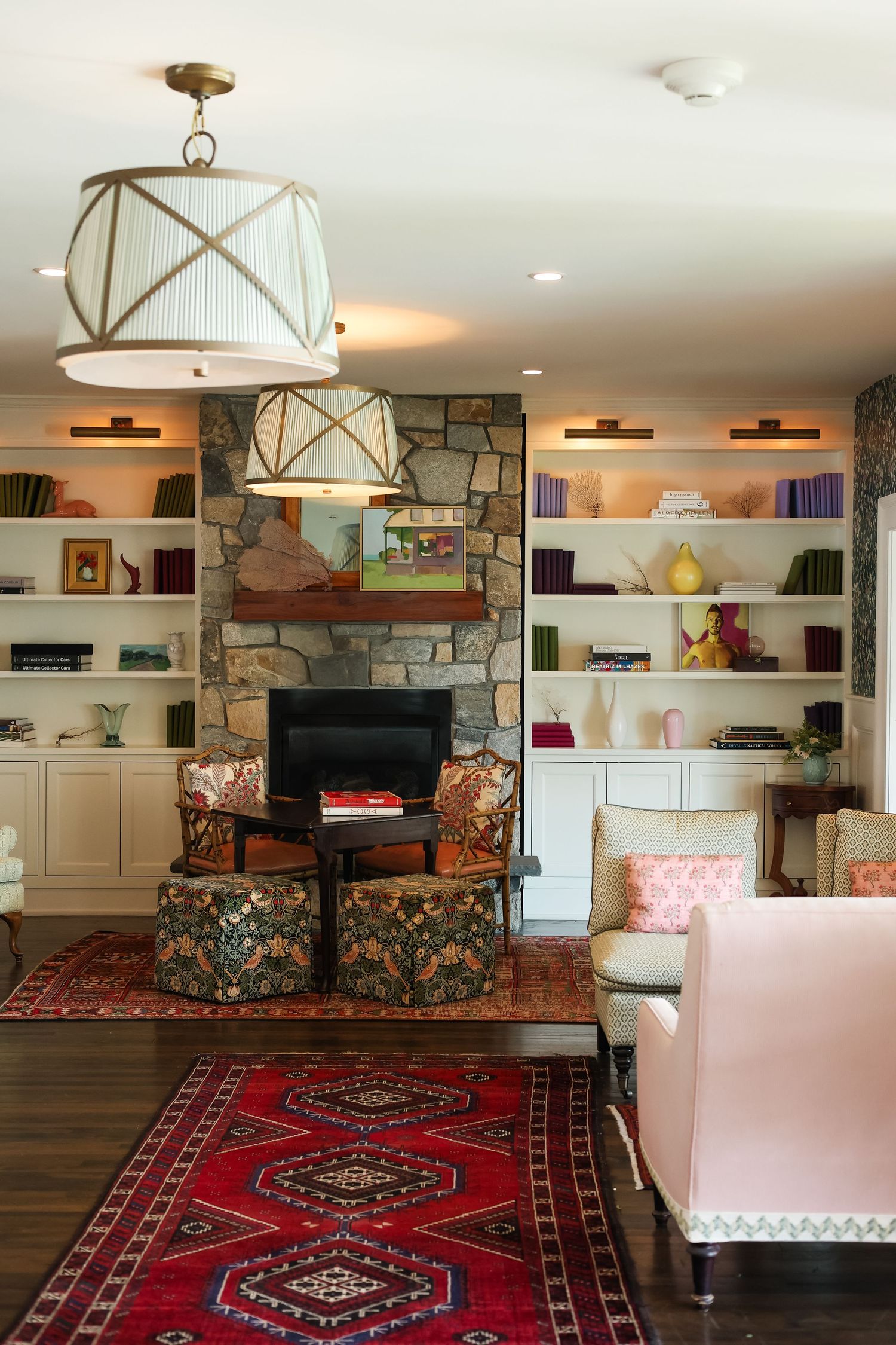 Cozy living room with stone fireplace, built-in shelves, floral chairs, red Persian rug, and elegant drum pendant light.