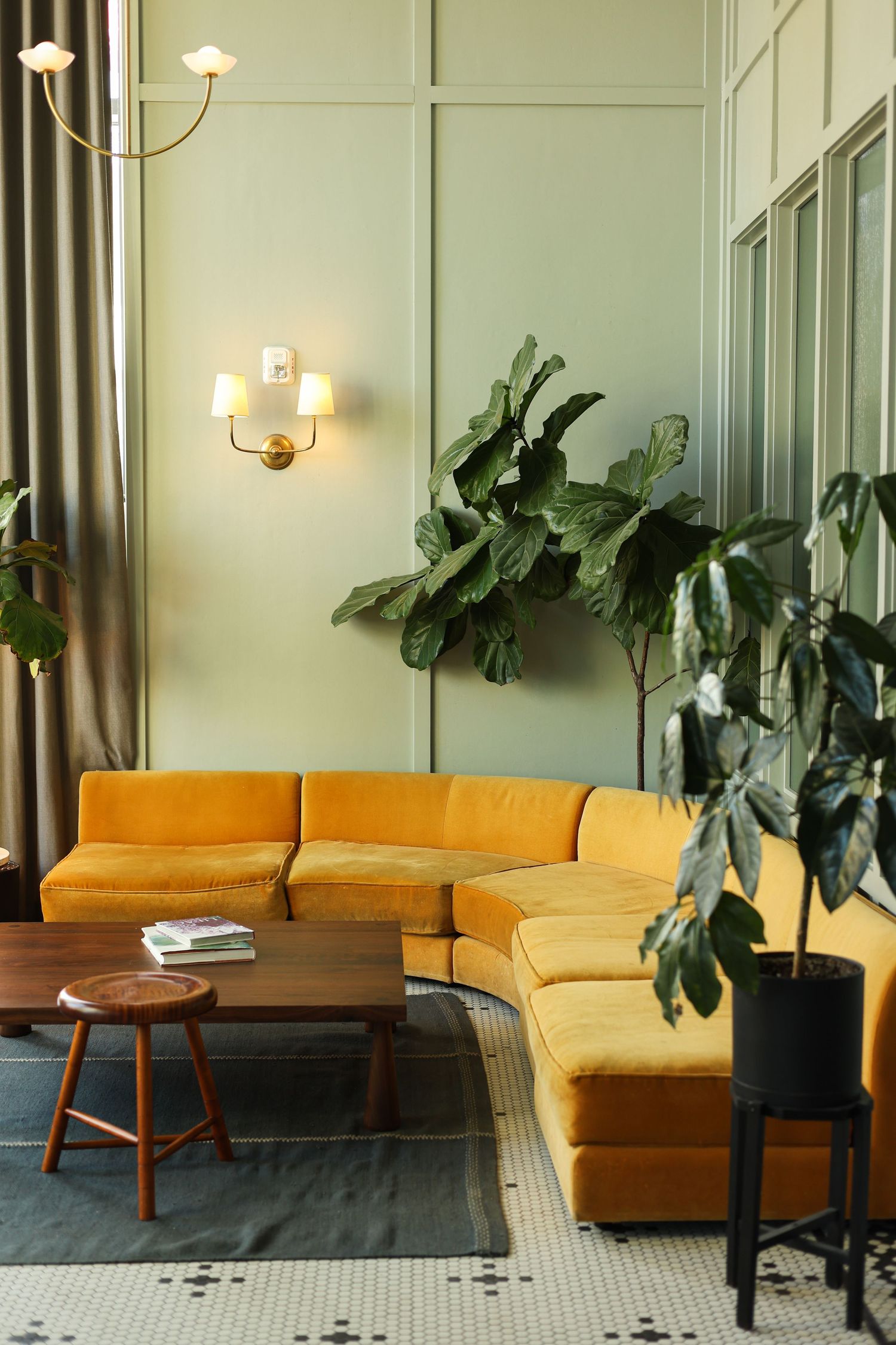Modern living room interior with yellow sectional sofa, wall sconces, and large potted plants against sage green walls.