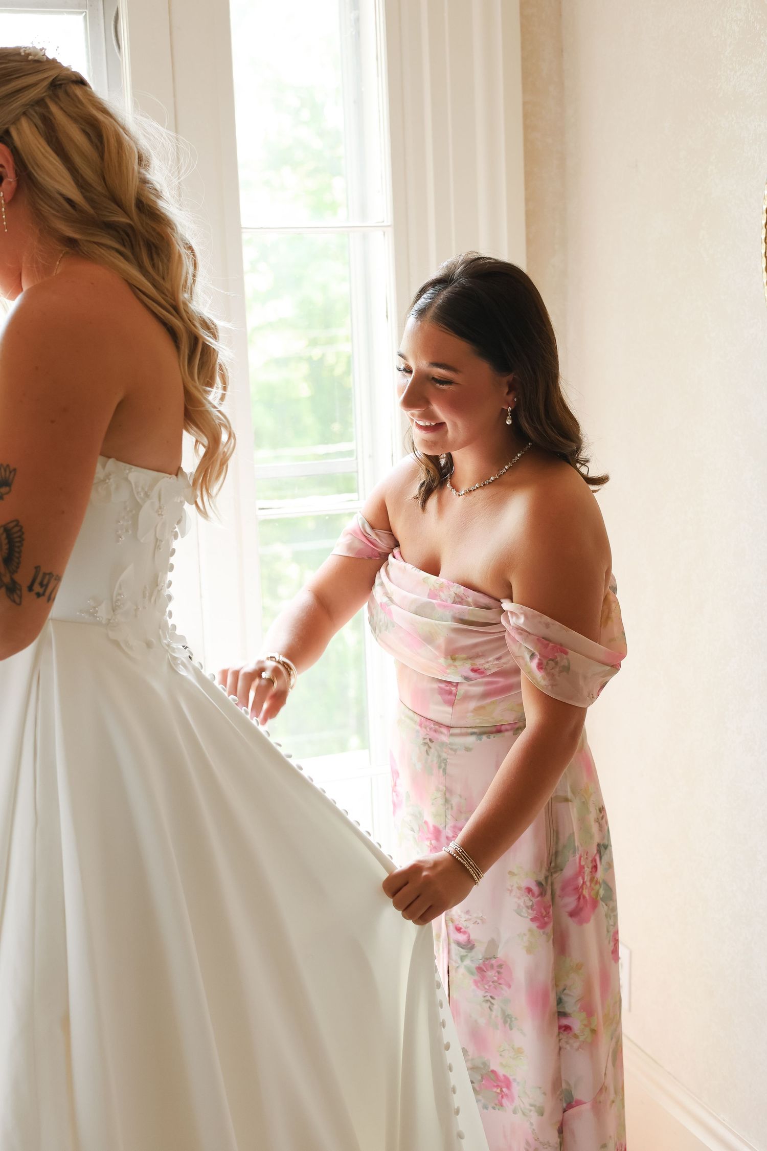 A smiling bridesmaid in a floral dress helps a bride in a white gown near a bright window.