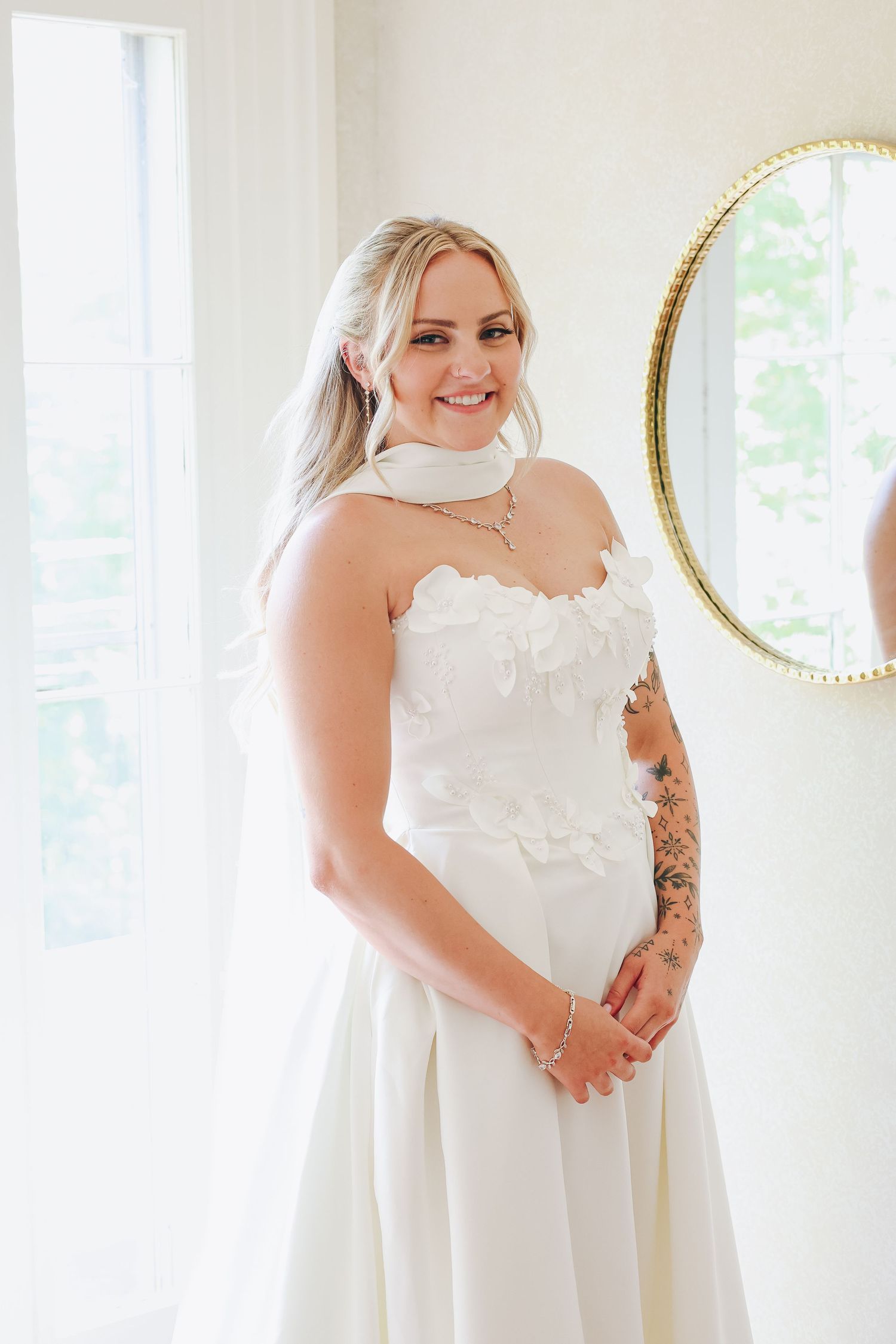 Smiling bride in strapless white floral wedding gown with tattoo, posing in bright room with gold mirror.