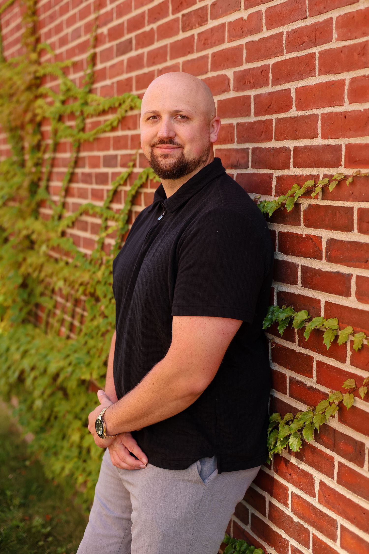 Bald man with beard in black polo shirt leaning against red brick wall with ivy, smiling confidently.