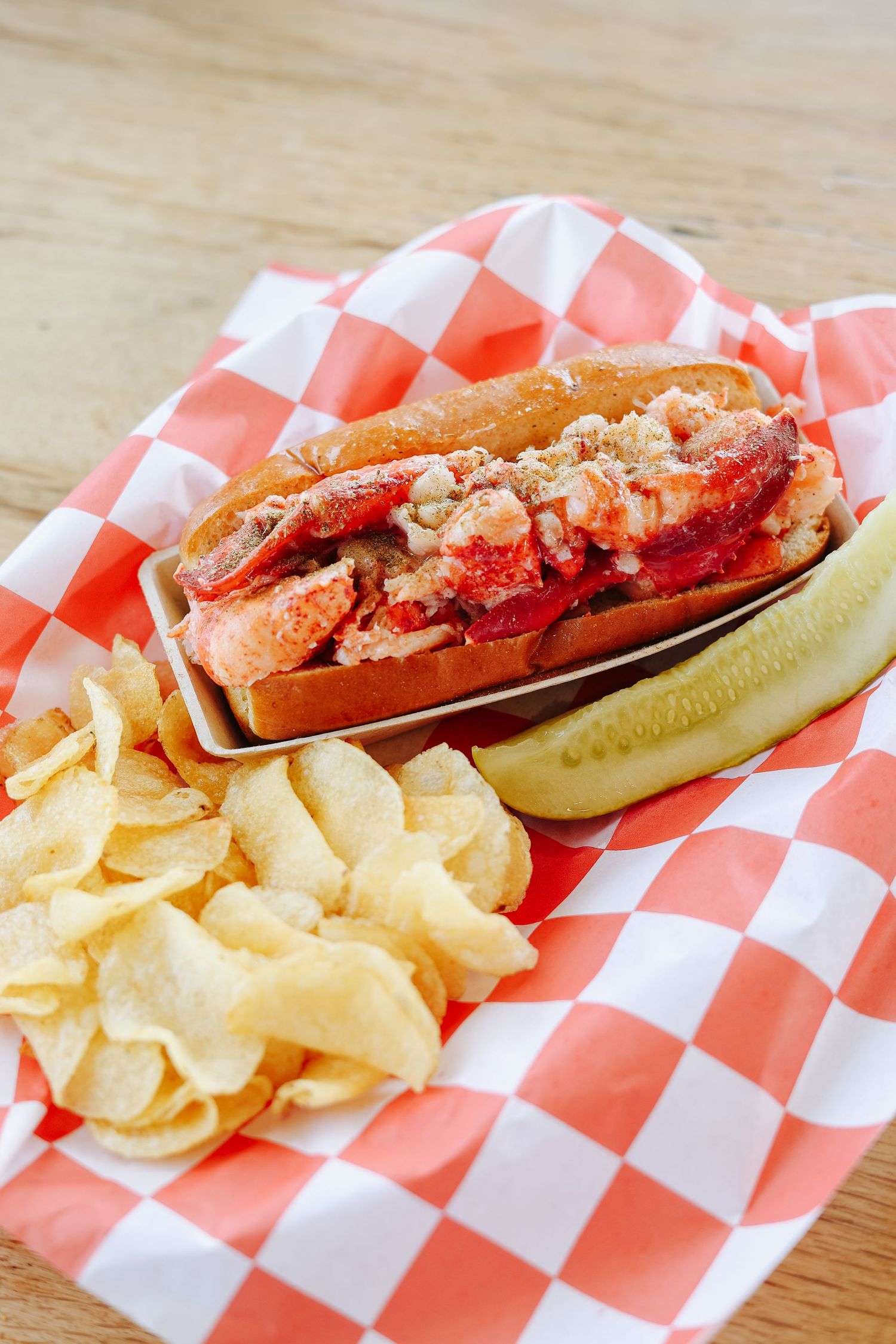 A classic lobster roll served in a toasted bun with chips and a pickle on red checkered paper.