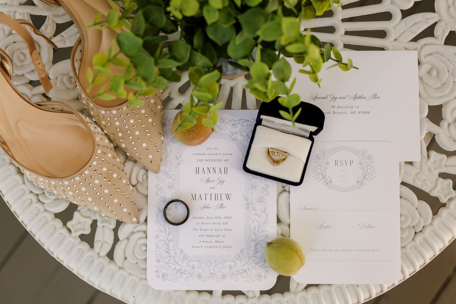 Wedding flat lay with invitation suite, gold rings, bridal heels, and greenery on white ornate table.