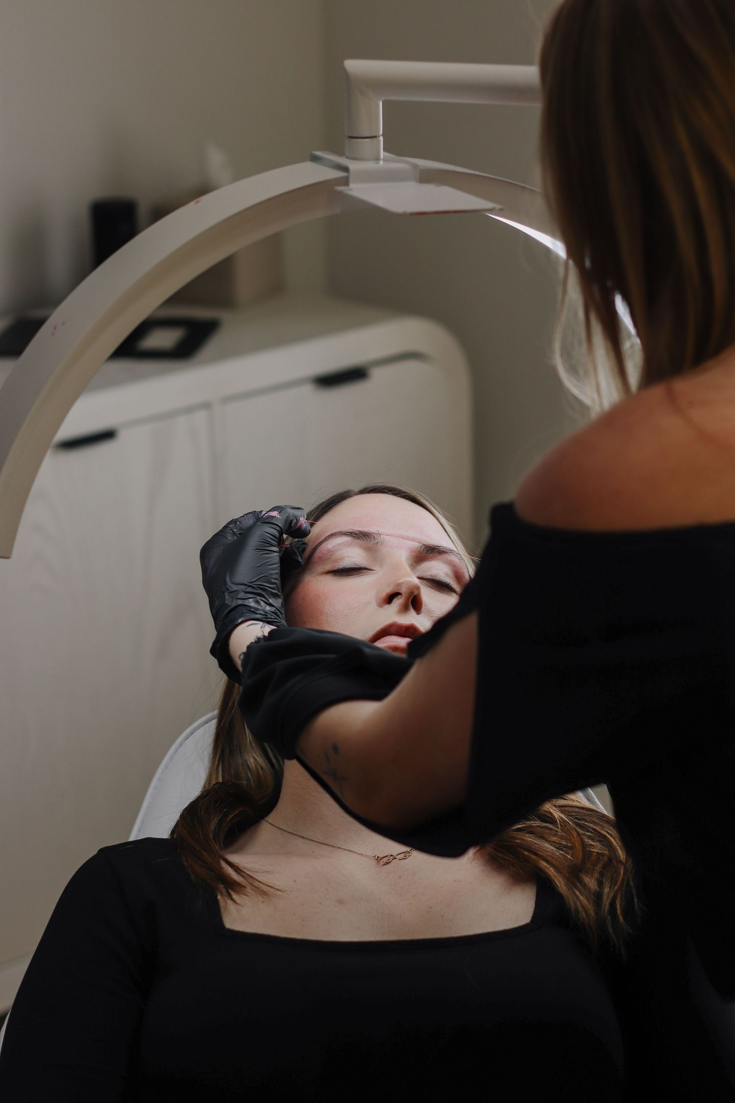 A client reclines in a salon chair during a professional brow waxing under bright overhead lighting.