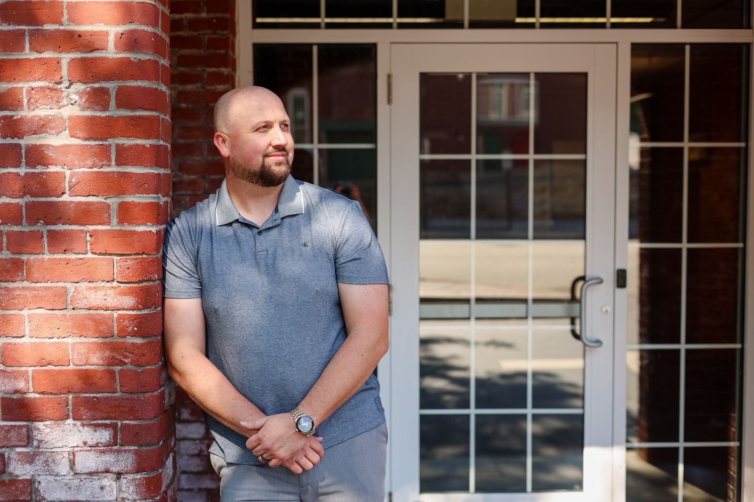 Man in gray polo shirt leaning against brick wall near glass door entrance outdoors.