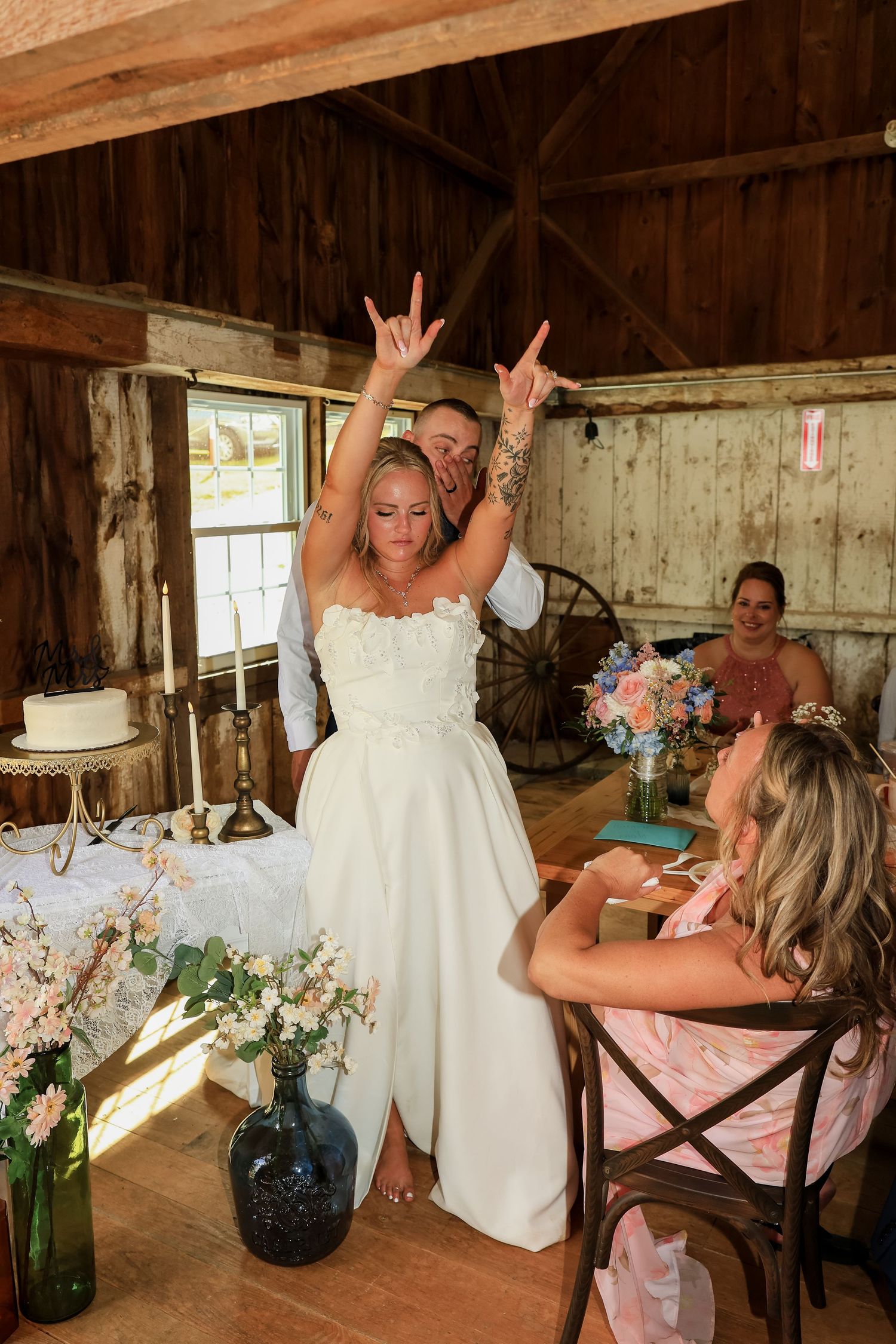Bride in white dress celebrates with hands raised at rustic barn wedding reception with guests.