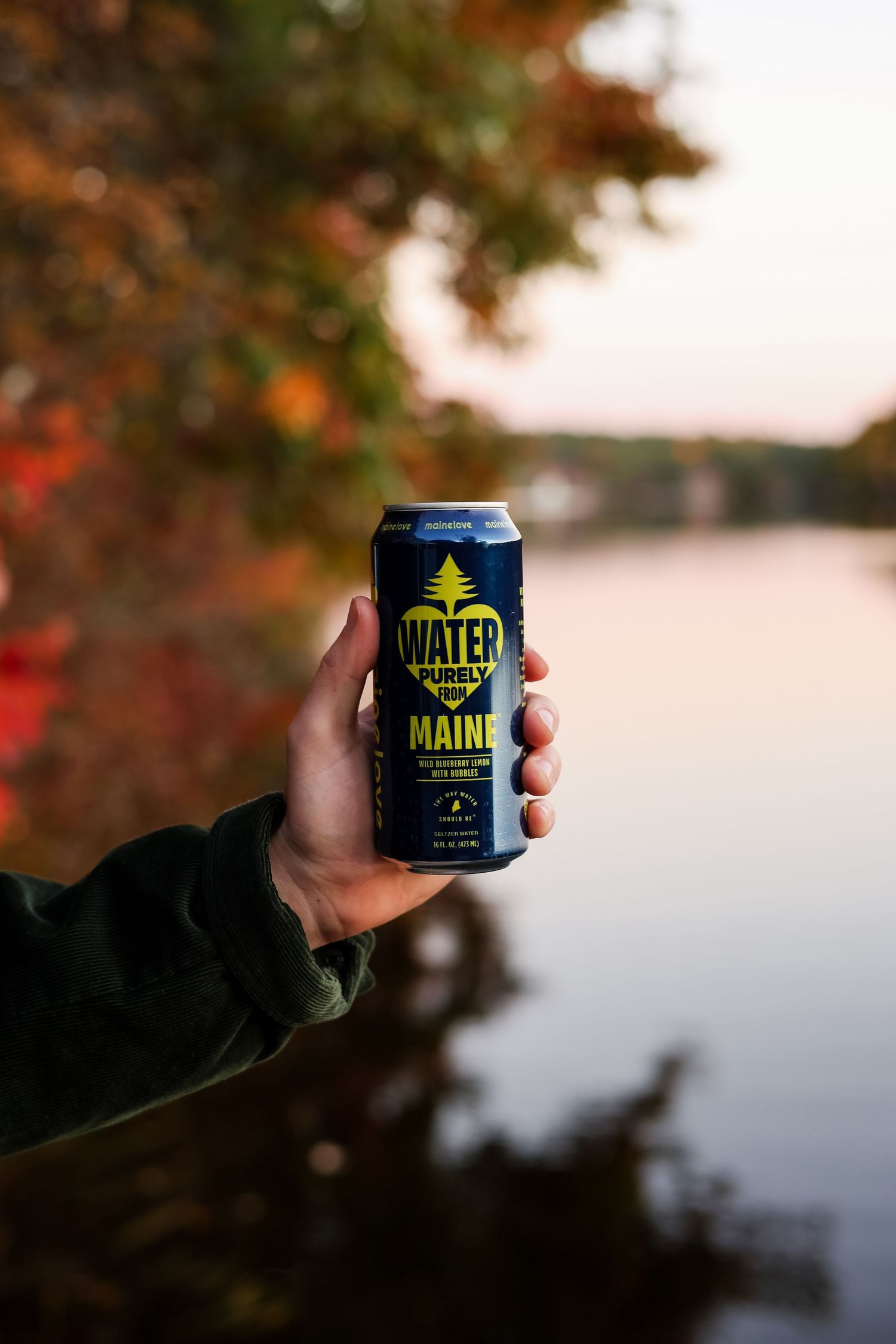 Hand holding a navy can of Water Purely from Maine beside a calm lake with colorful autumn foliage at dusk.