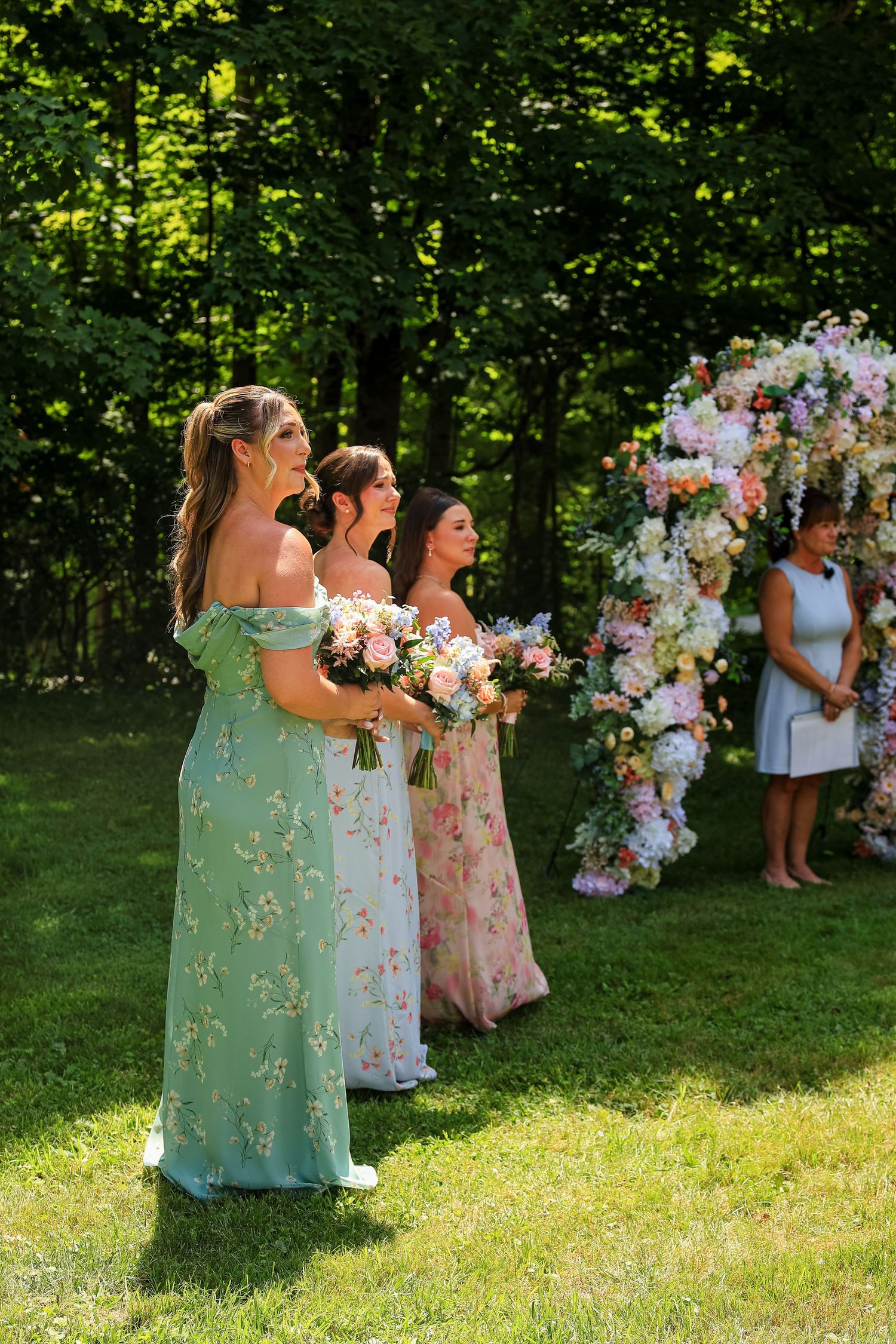 Bridesmaids in floral dresses holding bouquets stand outdoors near a flower arch at a garden wedding ceremony.