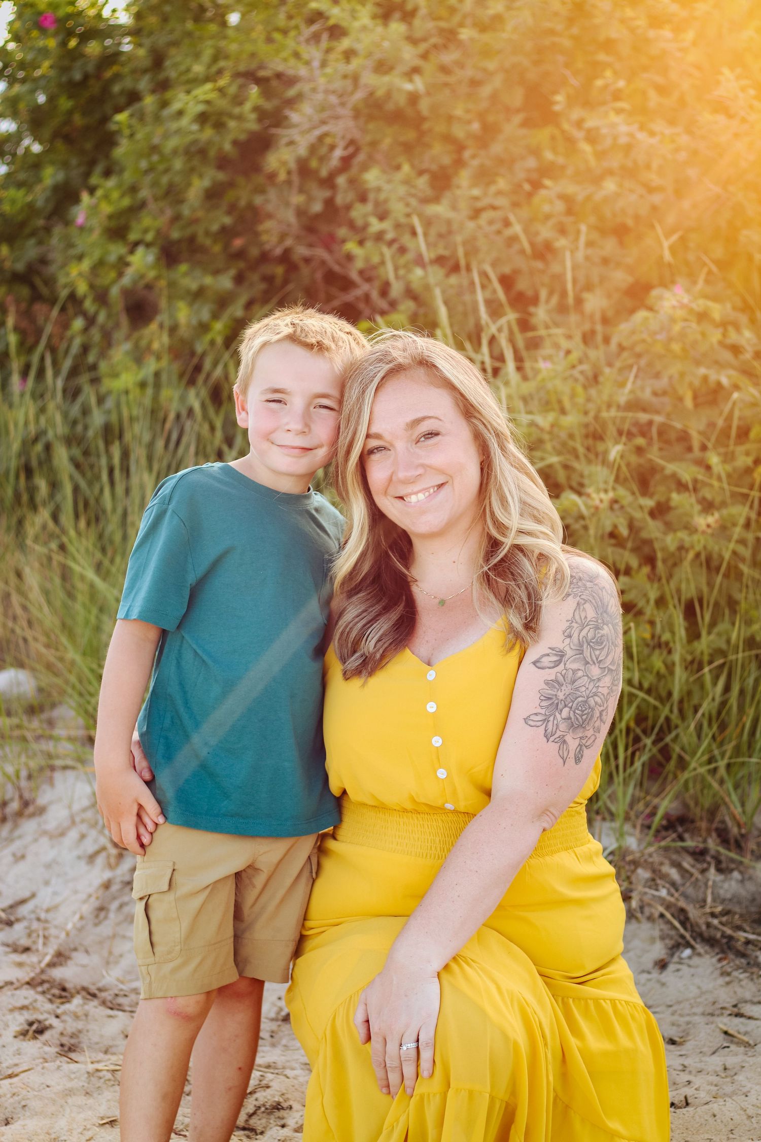 A smiling woman in a yellow dress with a floral tattoo poses with a young boy in a teal shirt on a sandy beach.