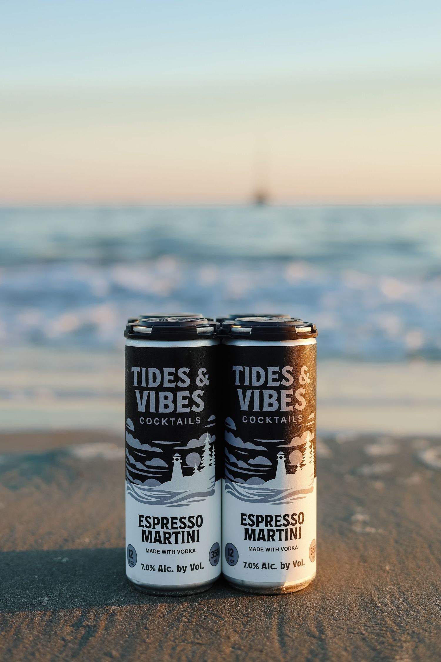 Two Tides & Vibes Espresso Martini vodka cocktail cans sit on a sandy beach with ocean waves and a calm sunset sky behind.