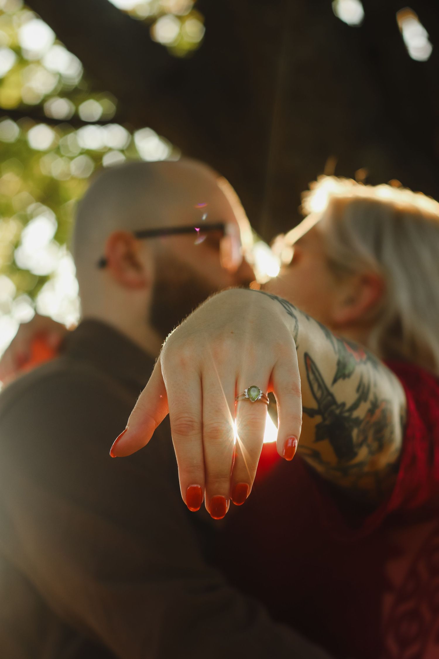 Couple kissing during engagement photo session, woman showing off diamond ring with tattoos and red nails in golden sunlight.