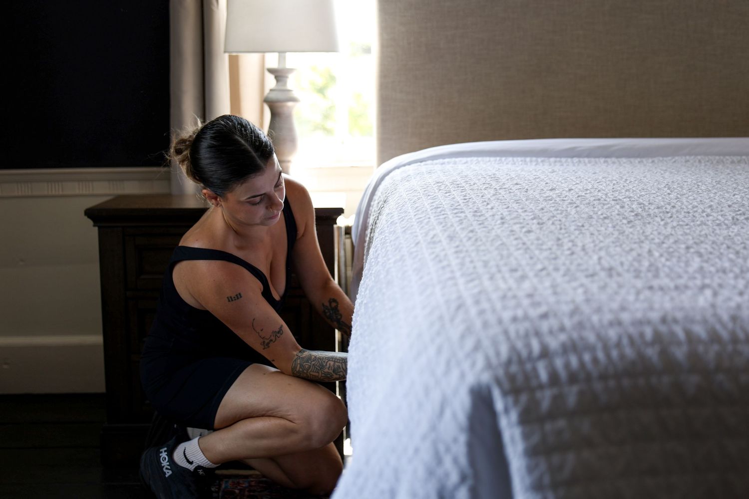 Person lying on a white bed in a dimly lit room with natural light coming through a window.