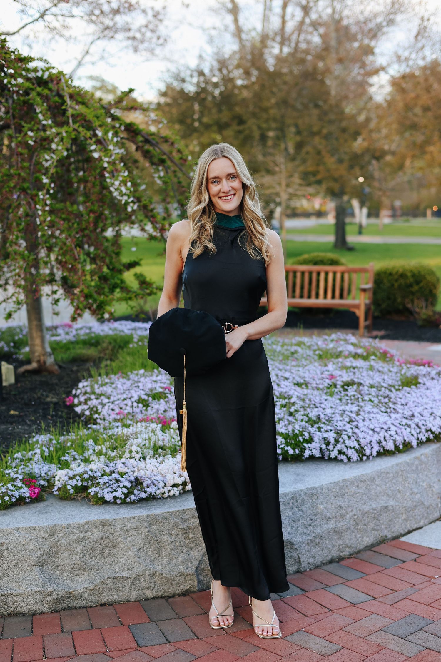 Woman in black sleeveless maxi dress holding a hat, standing in a garden with colorful flowers and a bench.