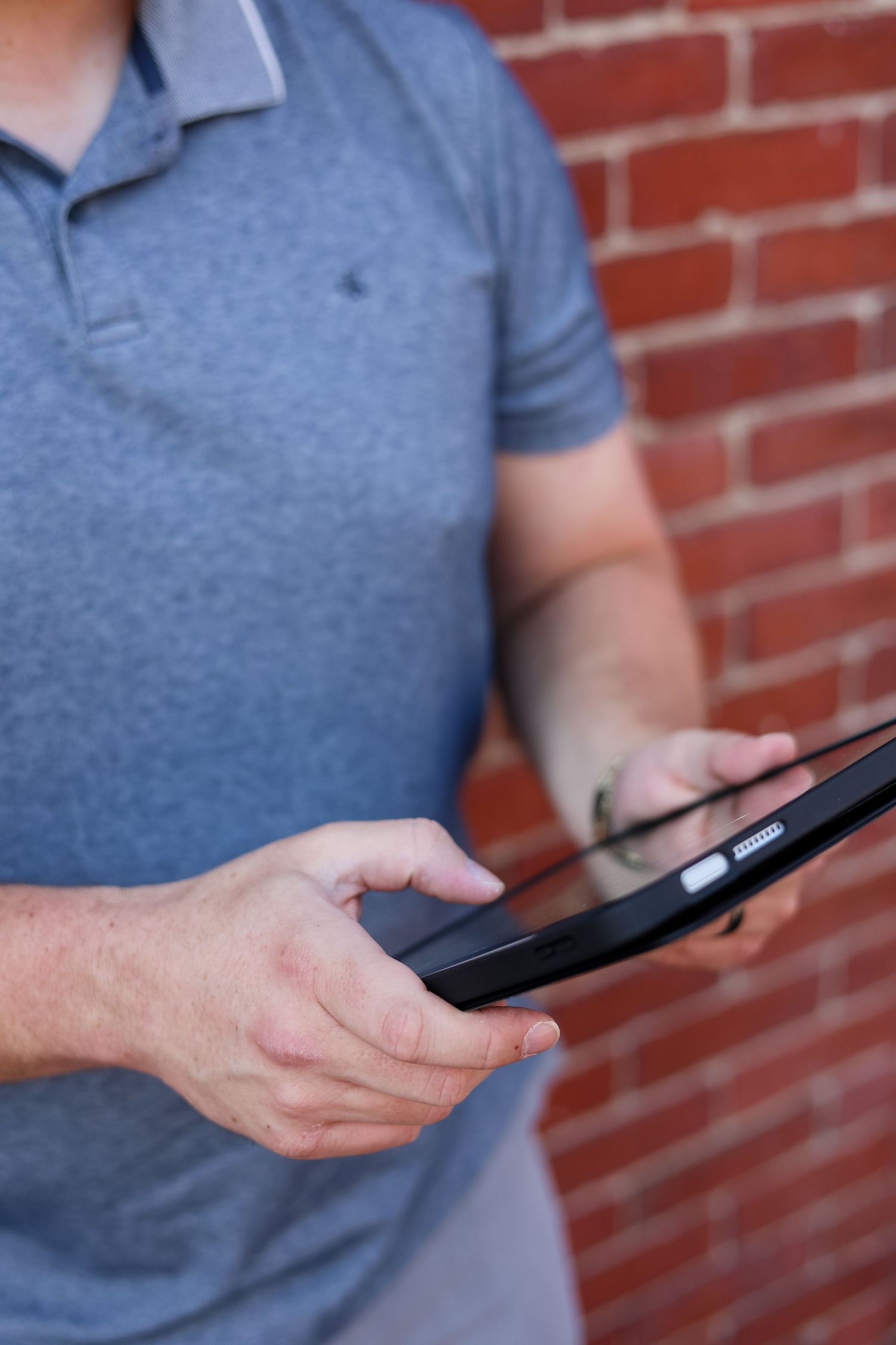 Man in gray polo shirt using a tablet computer against a red brick wall background.