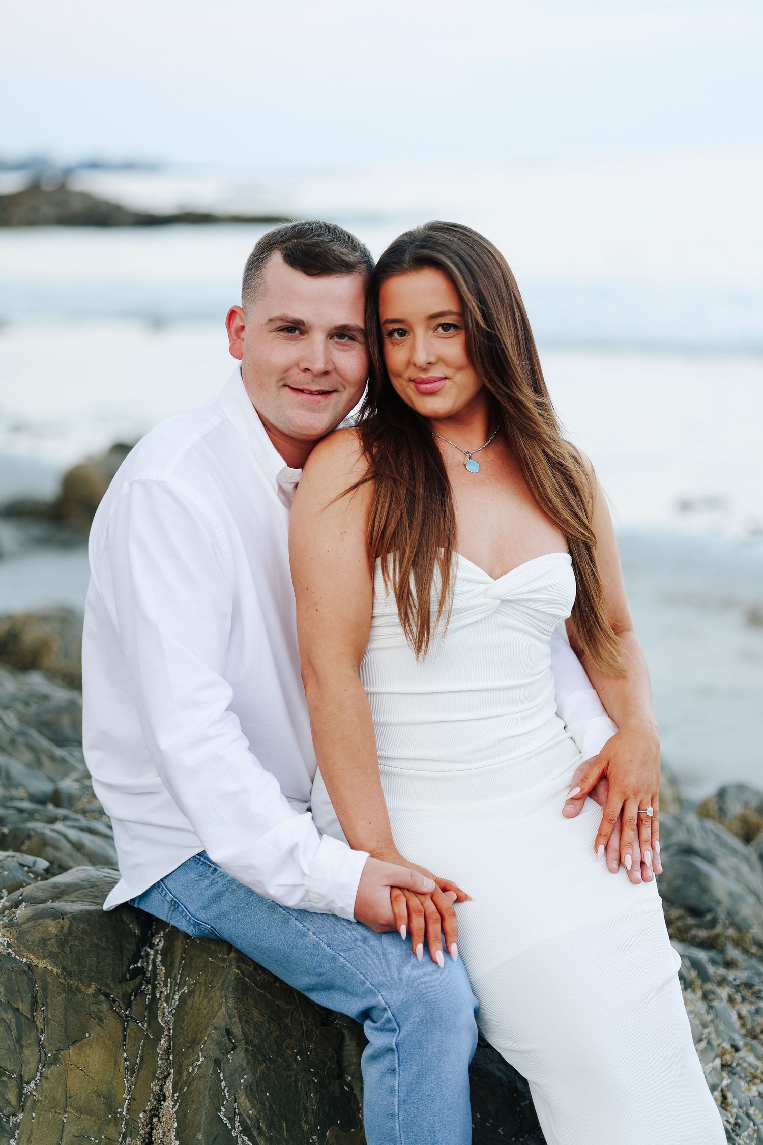 Romantic couple in white and denim embracing on rocky beach during sunset photoshoot.