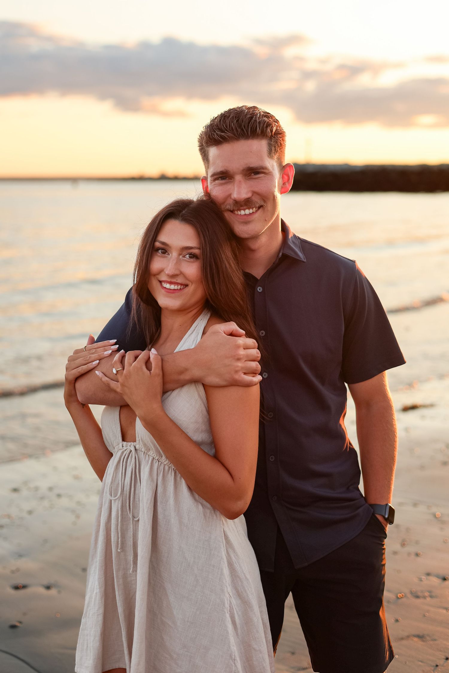 Couple embracing on a beach at sunset, smiling warmly, woman in white dress, man in navy shirt.