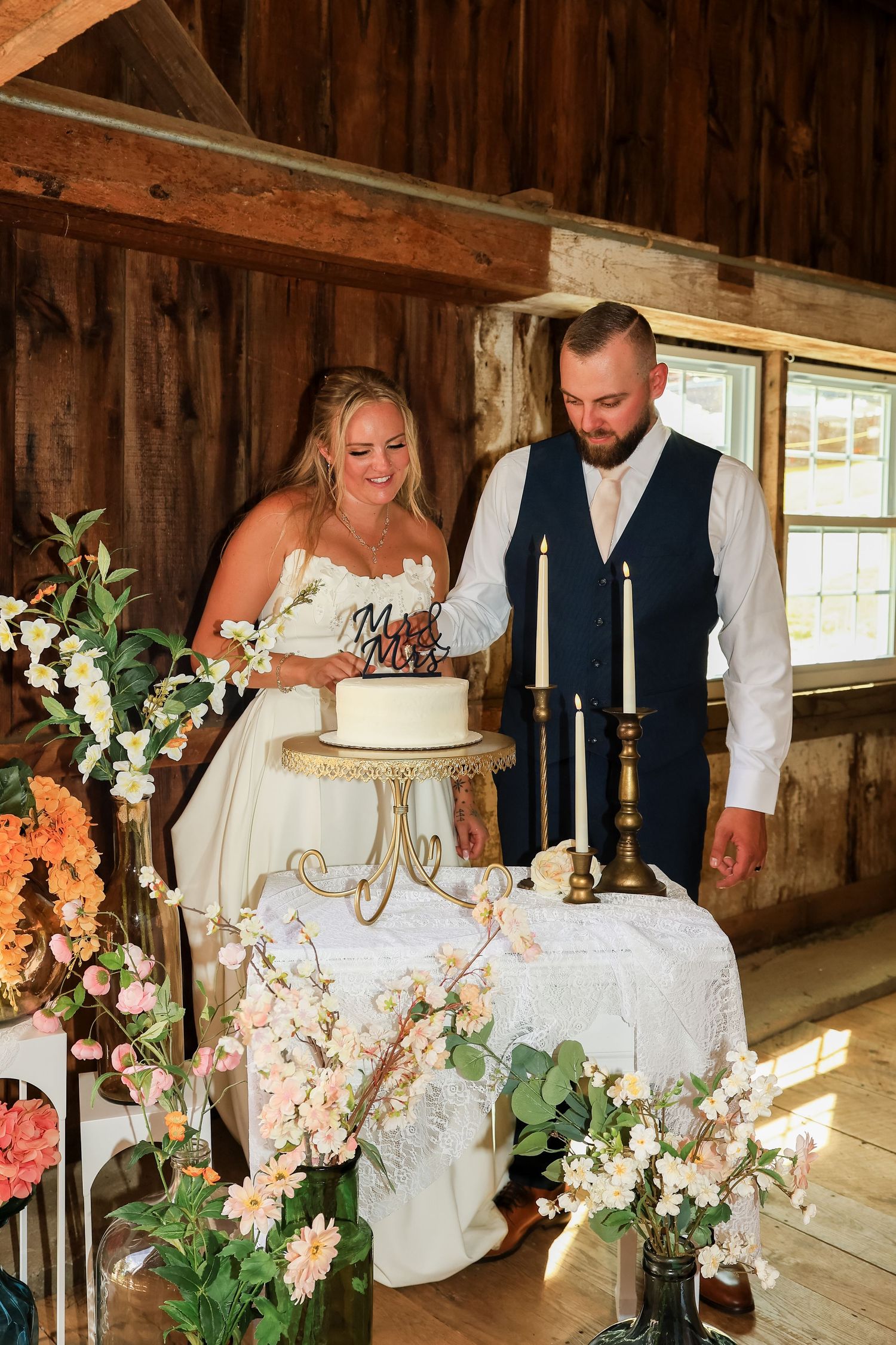 Bride and groom cutting white wedding cake in rustic barn venue surrounded by pink and white floral arrangements.