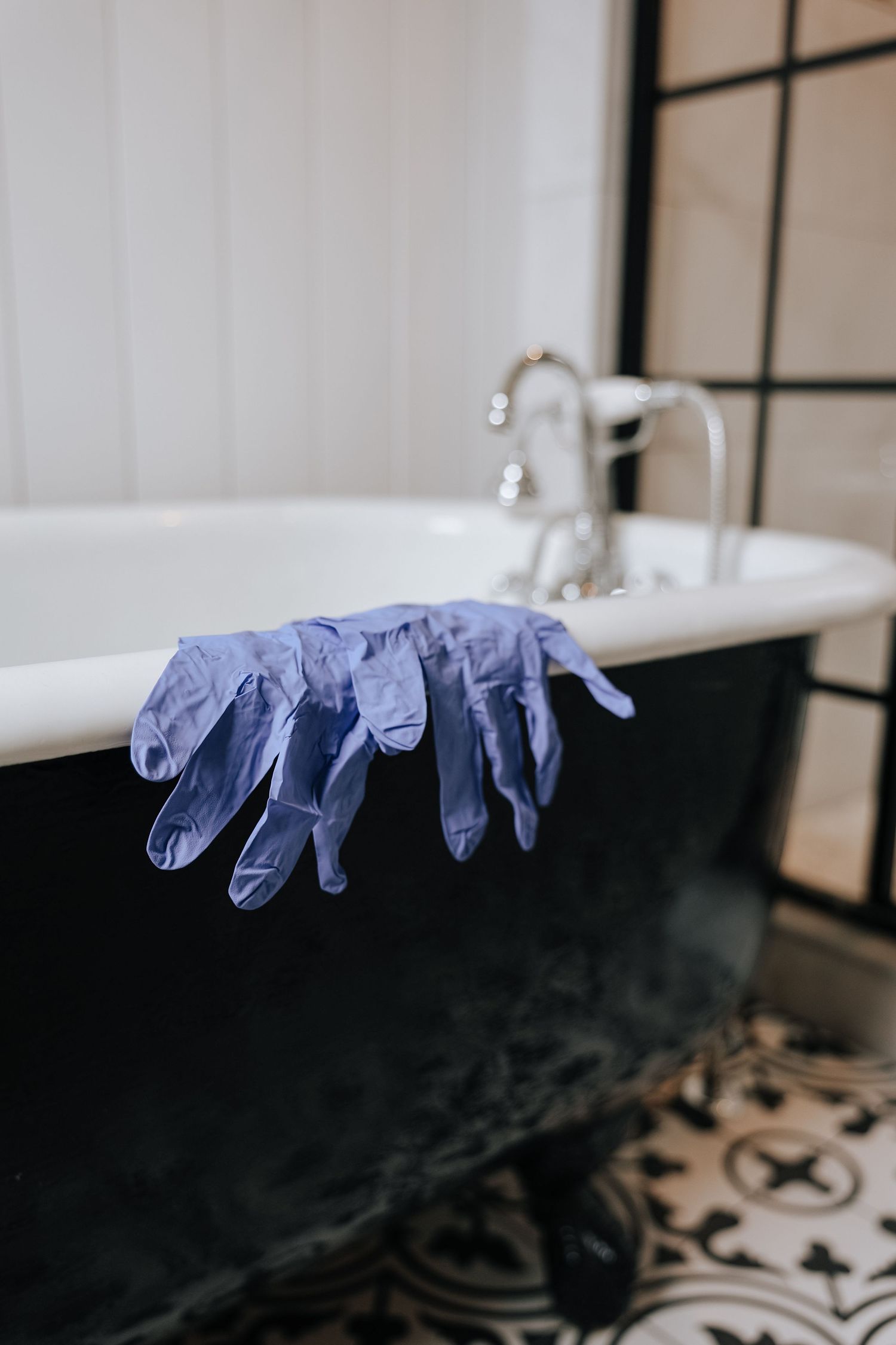 Purple latex gloves hang over the edge of a vintage black clawfoot bathtub with ornate tile flooring in bathroom.