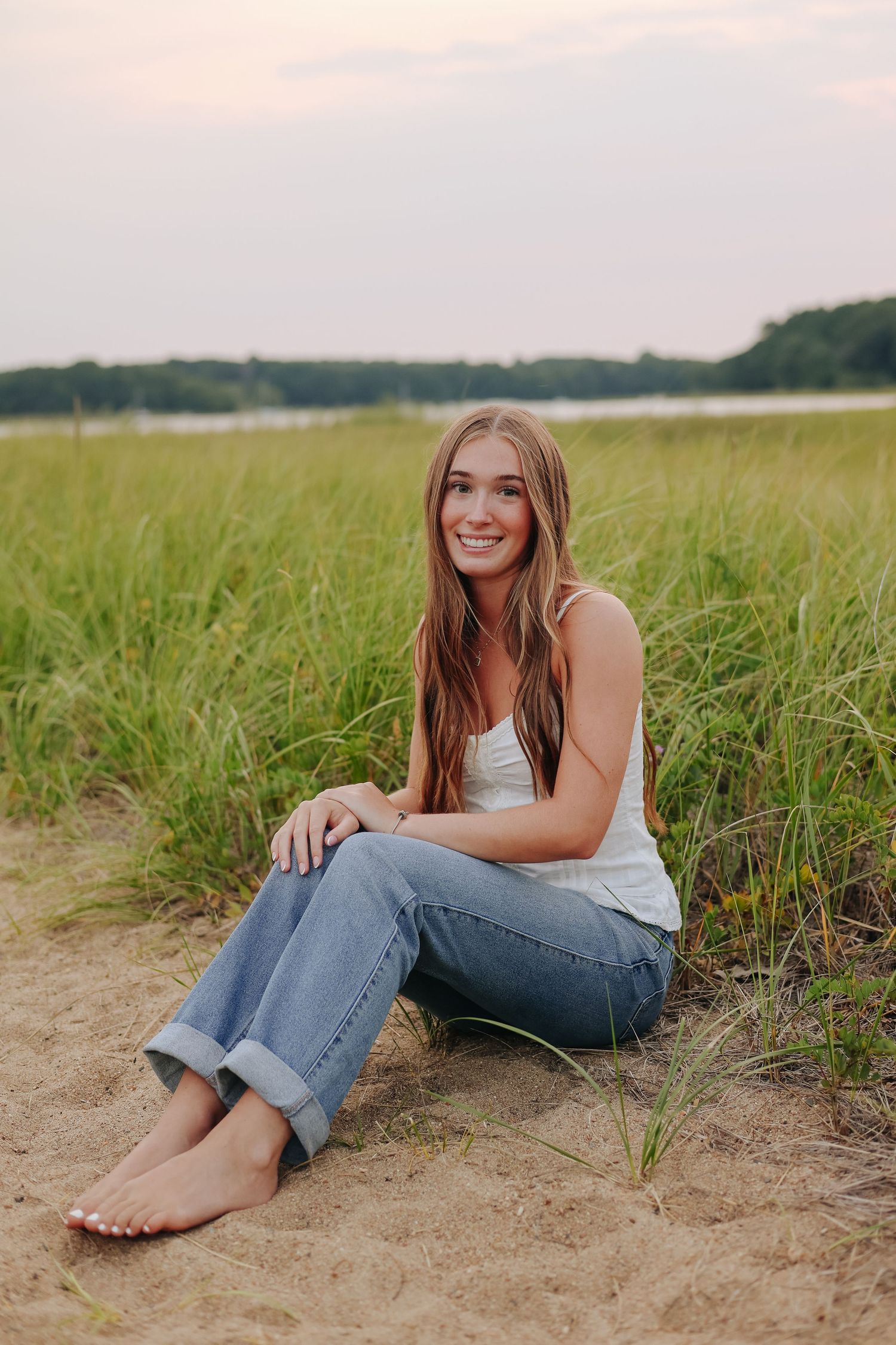 Smiling young woman sitting barefoot on sandy beach grass in jeans and white tank top at sunset.